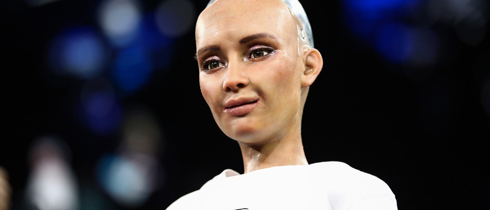 Ein humanoider Roboter mit einem realistischen Gesichtsausdruck steht im Vordergrund. Der Roboter tr&auml;gt ein wei&szlig;es Oberteil mit einem aufgedruckten Muster. Der Hintergrund ist unscharf und zeigt blaue und schwarze Farbt&ouml;ne. Der Roboter scheint in einer technologischen oder wissenschaftlichen Umgebung pr&auml;sentiert zu werden.