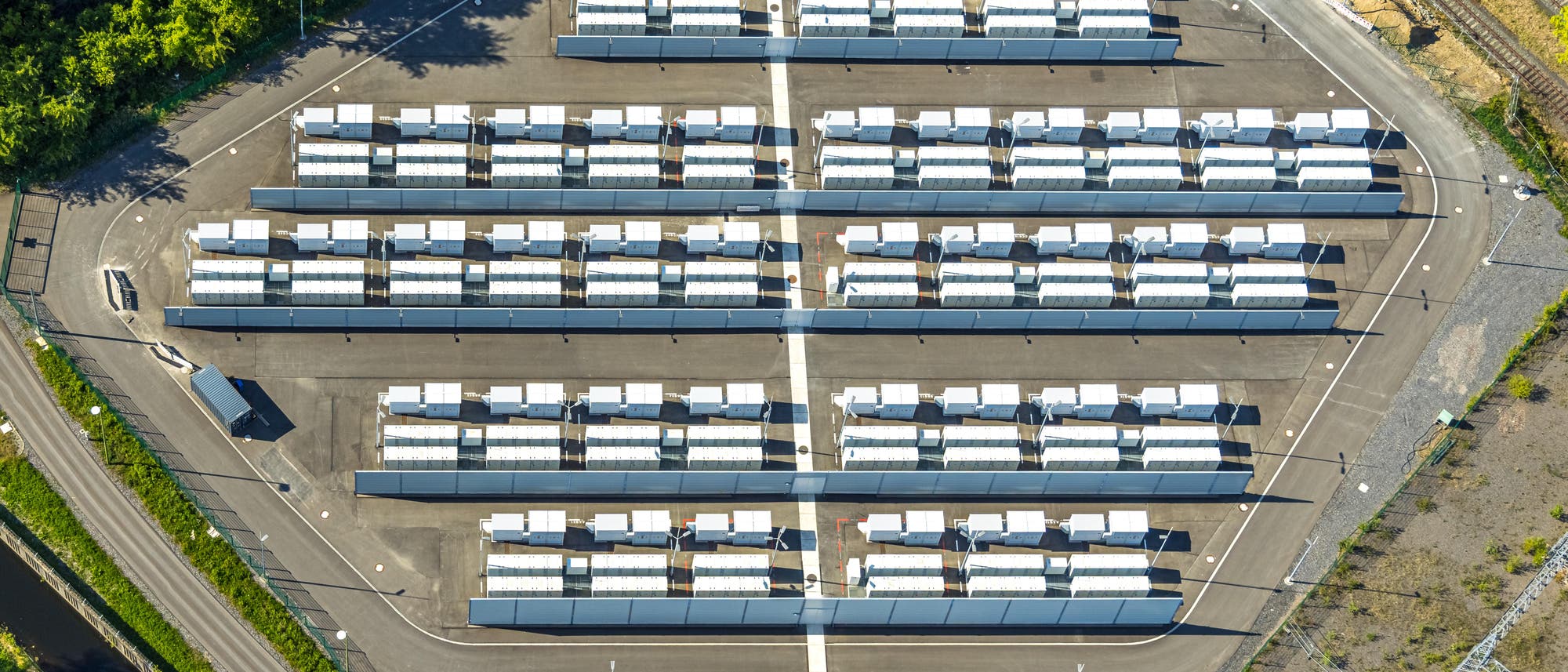 Luftaufnahme eines großen Solarkraftwerks mit mehreren Reihen von Solarmodulen, die auf einem asphaltierten Gelände angeordnet sind. Die Module sind in parallelen Linien aufgestellt, umgeben von Straßen und Vegetation. Im Vordergrund befindet sich ein Gebäude, das möglicherweise als Kontrollzentrum dient. Die Szene zeigt eine klare, strukturierte Anordnung der Solarpanels, die auf nachhaltige Energieerzeugung hinweist. Luftaufnahme eines großen Solarkraftwerks mit mehreren Reihen von Solarmodulen, die auf einem asphaltierten Gelände angeordnet sind. Die Module sind in parallelen Linien aufgestellt, umgeben von Straßen und Vegetation. Im Vordergrund befindet sich ein Gebäude, das möglicherweise als Kontrollzentrum dient. Die Szene zeigt eine klare, strukturierte Anordnung der Solarpanels, die auf nachhaltige Energieerzeugung hinweist.