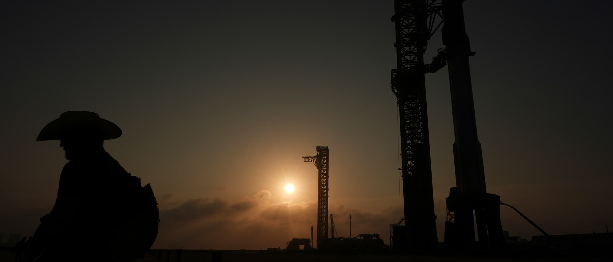 Ein Sonnenuntergang mit der Silhouette eines Mannes mit Cowboyhut und Rucksack. Rechts im Bild sind zwei gro&szlig;e, turmartige Strukturen einer Raketenabschussrampe zu sehen. 