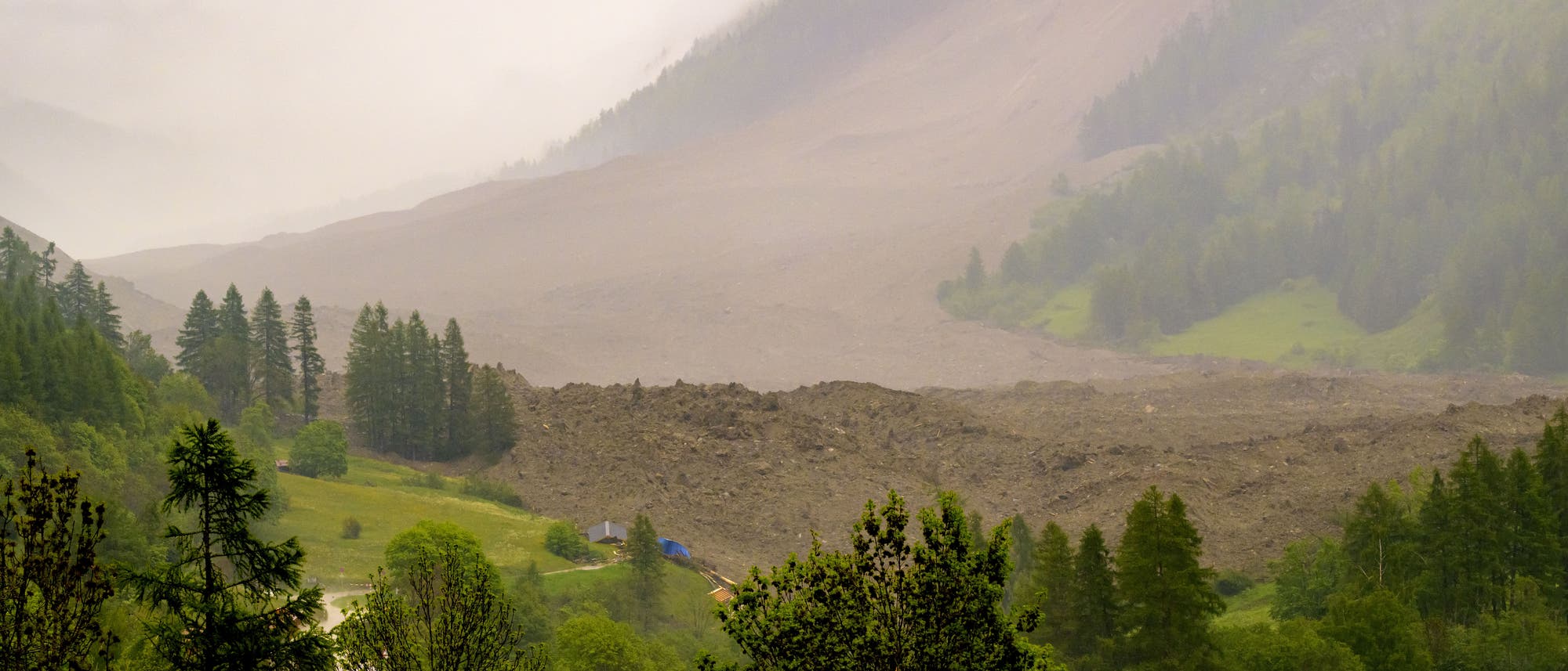 Eine weitläufige Landschaft mit einem großen Erdrutsch, der einen Hang hinunterfließt. Im Vordergrund sind grüne Bäume und Wiesen zu sehen, während der Himmel bewölkt ist und Nebel die Berge umhüllt. Keine Menschen und nur wenige Gebäude sind sichtbar. Eine weitläufige Landschaft mit einem großen Erdrutsch, der einen Hang hinunterfließt. Im Vordergrund sind grüne Bäume und Wiesen zu sehen, während der Himmel bewölkt ist und Nebel die Berge umhüllt. Keine Menschen und nur wenige Gebäude sind sichtbar.