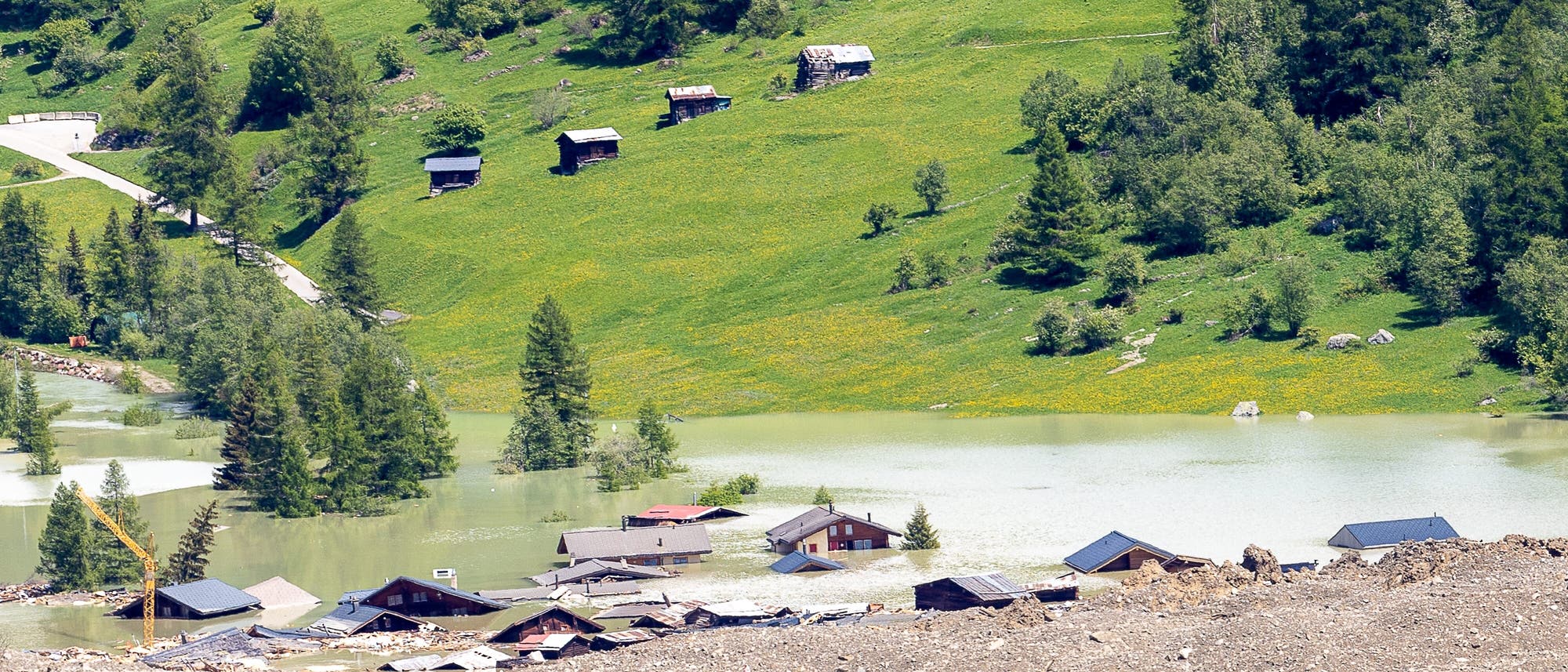 Eine ländliche Landschaft in den Schweizer Alpen mit grünen Hügeln und vereinzelten Bäumen. Im Vordergrund sind mehrere Gebäude teilweise von Hochwasser überflutet. Die Dächer der Häuser ragen aus dem Wasser hervor. Im Hintergrund sind kleine Holzhütten auf einer grünen Wiese zu sehen. Ein Kran ist links im Bild sichtbar. 