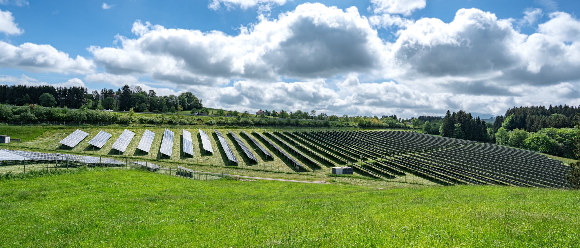 Eine weitläufige Solaranlage auf einem grünen Hügel unter einem bewölkten Himmel. Die Solarpaneele sind in geordneten Reihen angeordnet und erstrecken sich über die Landschaft. Im Hintergrund sind Bäume und ein paar Gebäude zu sehen. Die Szene vermittelt ein Gefühl von nachhaltiger Energieerzeugung und Umweltschutz. Eine weitläufige Solaranlage auf einem grünen Hügel unter einem bewölkten Himmel. Die Solarpaneele sind in geordneten Reihen angeordnet und erstrecken sich über die Landschaft. Im Hintergrund sind Bäume und ein paar Gebäude zu sehen. Die Szene vermittelt ein Gefühl von nachhaltiger Energieerzeugung und Umweltschutz.