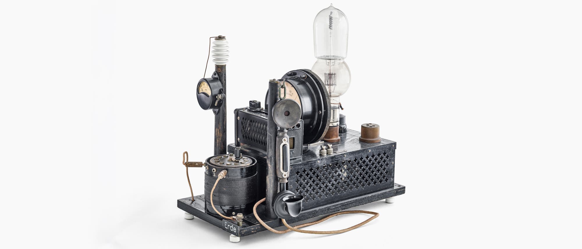 Ein altes elektrisches Ger&auml;t mit verschiedenen Komponenten, darunter die Liebenr&ouml;hre, die einer gro&szlig;e Gl&uuml;hbirne gleicht, Frequenzanzeigen und mehrere Kabel. Das Ger&auml;t steht auf einer rechteckigen Basis mit einem Gittermuster. Ein Schild mit der Aufschrift &bdquo;Erde&ldquo; ist an der Vorderseite angebracht. Das Design deutet auf eine historische technische Apparatur aus der fr&uuml;hen Elektronik- oder Funktechnik hin.