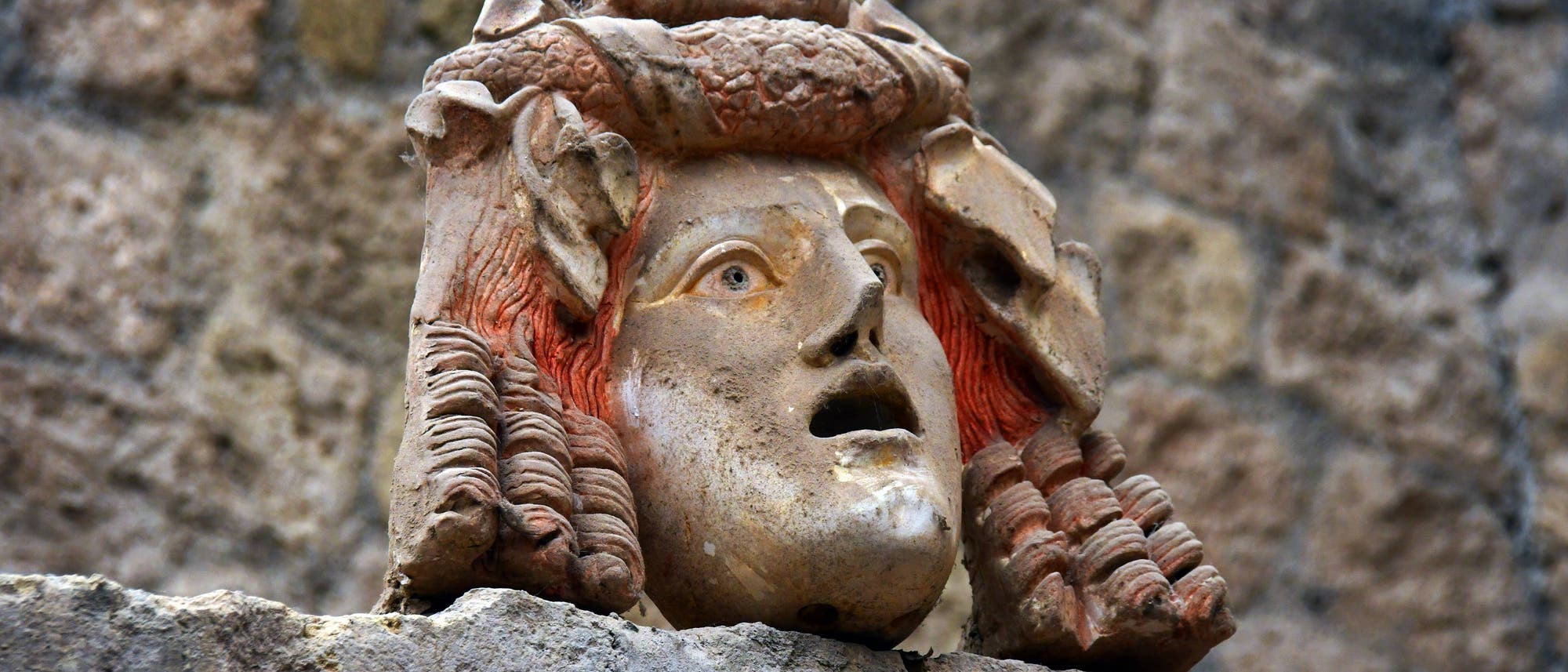 Steinerne Skulptur eines Kopfes mit ausdrucksstarkem Gesicht, roten Haaren und einem kunstvoll verzierten Kopfschmuck, der Früchte darstellt. Die Skulptur ist auf einer Steinmauer montiert, die als Hintergrund dient.