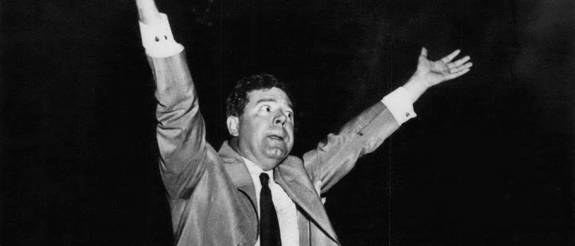 Ein Mann in einem Anzug hebt beide Arme in die Luft, als ob er eine Rede hält oder jubelt. Der Hintergrund ist dunkel, was den Fokus auf seine Gestik und Mimik lenkt.