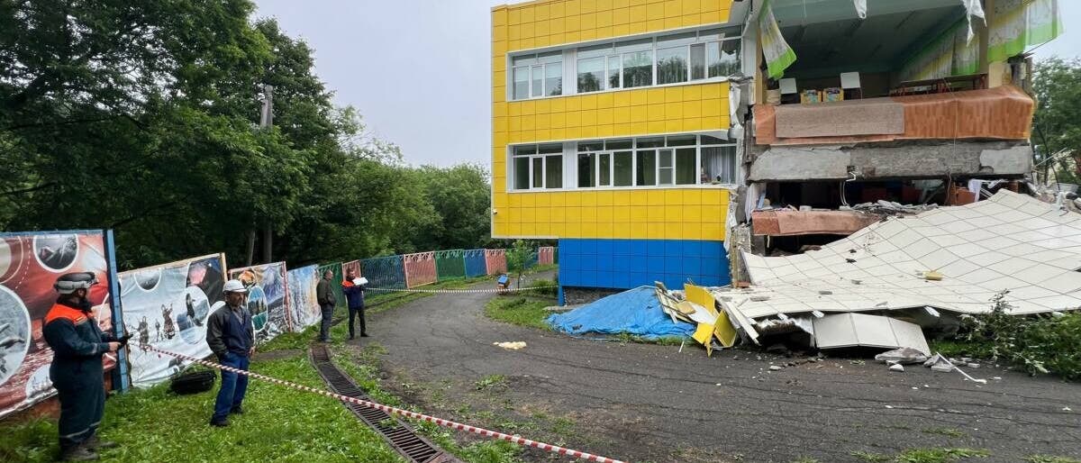 Ein teilweise eingestürztes Gebäude mit gelber und blauer Fassade. Der rechte Teil des Gebäudes ist stark beschädigt, mit herabgefallenen Trümmern und freiliegenden Innenräumen. Im Vordergrund stehen mehrere Personen in Schutzkleidung hinter einer Absperrung. Links sind Bäume und ein Zaun mit Plakaten zu sehen. Der Himmel ist bewölkt.