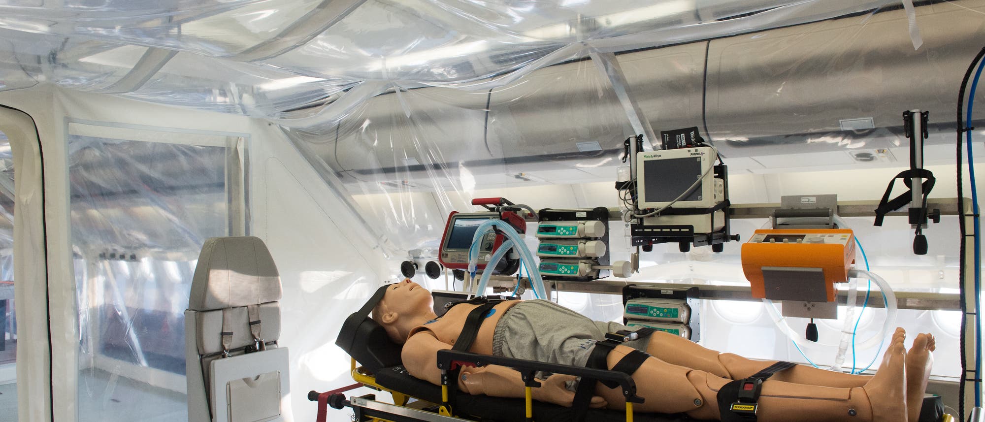 Eine medizinische Simulationseinrichtung mit einer lebensgro&szlig;en Puppe auf einer Trage, umgeben von medizinischen Ger&auml;ten wie Monitoren und Beatmungsger&auml;ten. Die Umgebung ist in einem transparenten Zelt aufgebaut, das m&ouml;glicherweise f&uuml;r Isolations- oder Trainingszwecke genutzt wird. Die Szene vermittelt den Eindruck einer realistischen Notfallsituation.