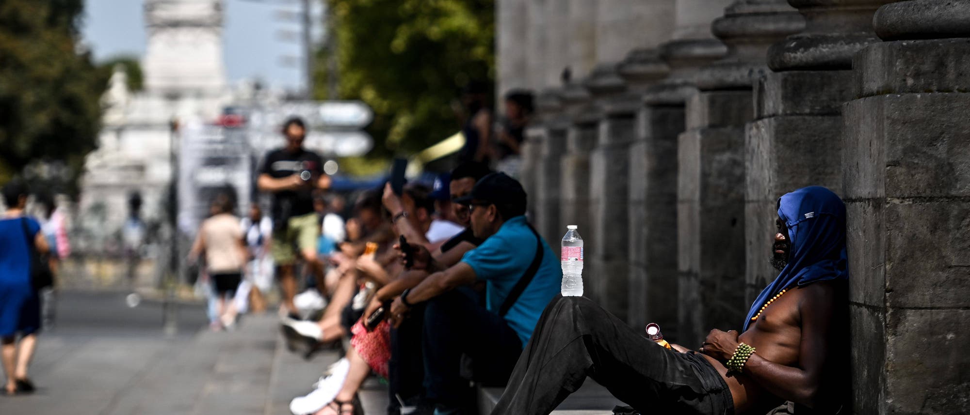 Eine Gruppe von Menschen sitzt auf den Stufen eines großen Gebäudes im Freien. Eine Person im Vordergrund lehnt sich mit einer Wasserflasche auf dem Bein zurück. Die Szene zeigt einen sonnigen, heißen Tag in einer städtischen Umgebung mit Bäumen und einer Statue im Hintergrund. Die Menschen scheinen sich auszuruhen oder zu erholen.
