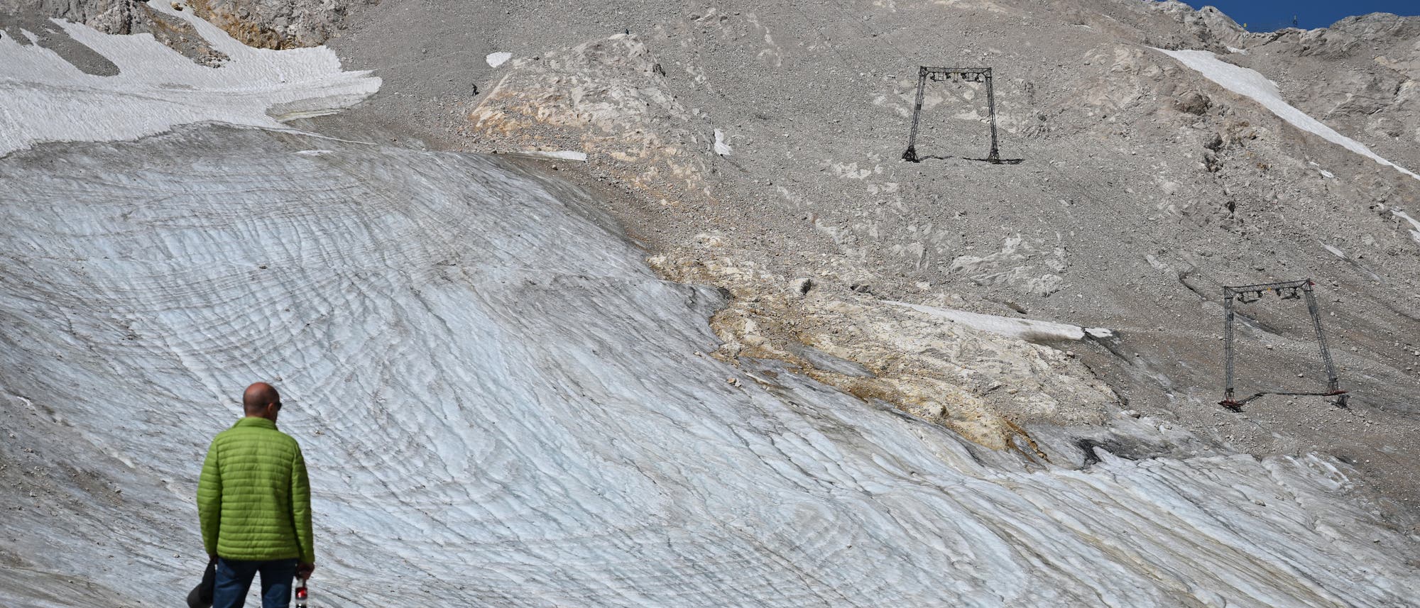 Eine Person in einer gr&uuml;nen Jacke steht vor einem Gletscher auf einem felsigen Berg. Der Gletscher zeigt deutliche Schmelzspuren und ist von Ger&ouml;ll umgeben. Im Hintergrund sind &Uuml;berreste von Skiliftstrukturen zu sehen. Der Himmel ist klar und blau.