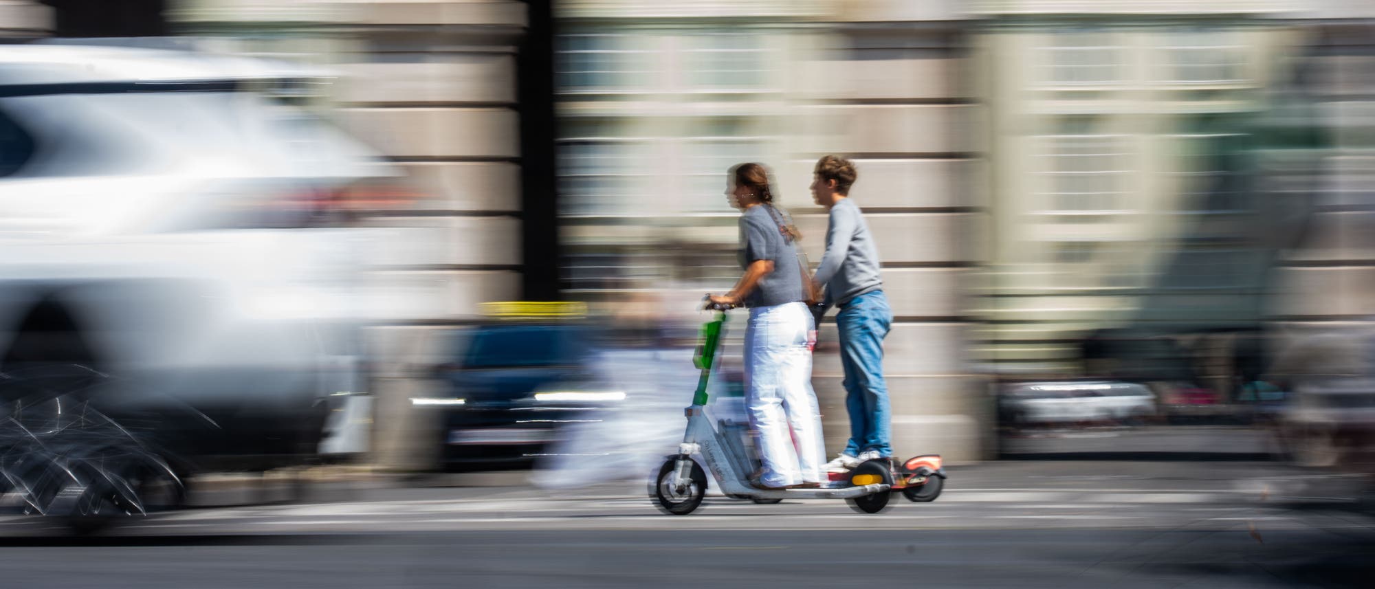 Zwei Personen fahren gemeinsam auf einem E-Scooter durch eine belebte Stadtstraße. Die Umgebung ist verschwommen, was die Bewegung und Geschwindigkeit betont. Im Hintergrund sind Gebäude und vorbeifahrende Autos zu erkennen.