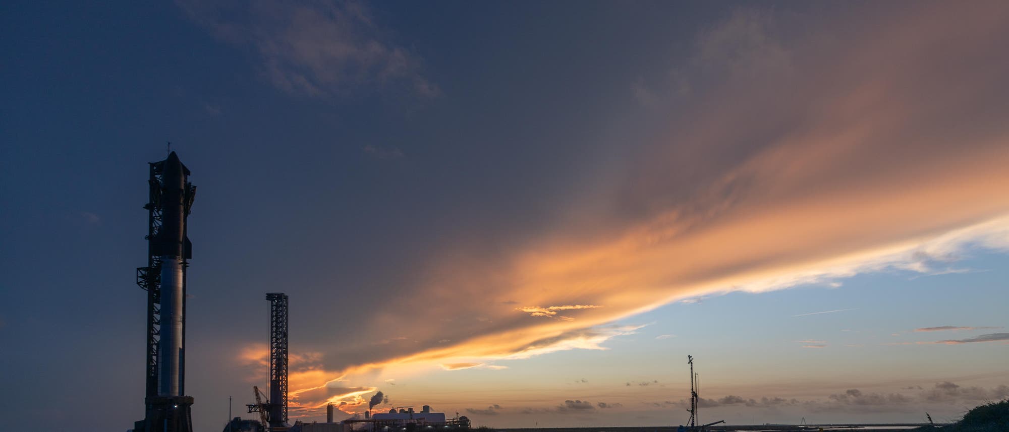 Un cohete está en una rampa inicial al atardecer. El cielo está coloreado con tonos de naranja y azul calientes, mientras que las nubes cruzan el horizonte. La escena transmite un ambiente tranquilo y de espera. En primer plano, la silueta de cohetes es claramente visible, rodeada de estructuras técnicas. Un cohete está en una rampa inicial al atardecer. El cielo está coloreado con tonos de naranja y azul calientes, mientras que las nubes cruzan el horizonte. La escena transmite un ambiente tranquilo y de espera. En primer plano, la silueta de cohetes es claramente visible, rodeada de estructuras técnicas.