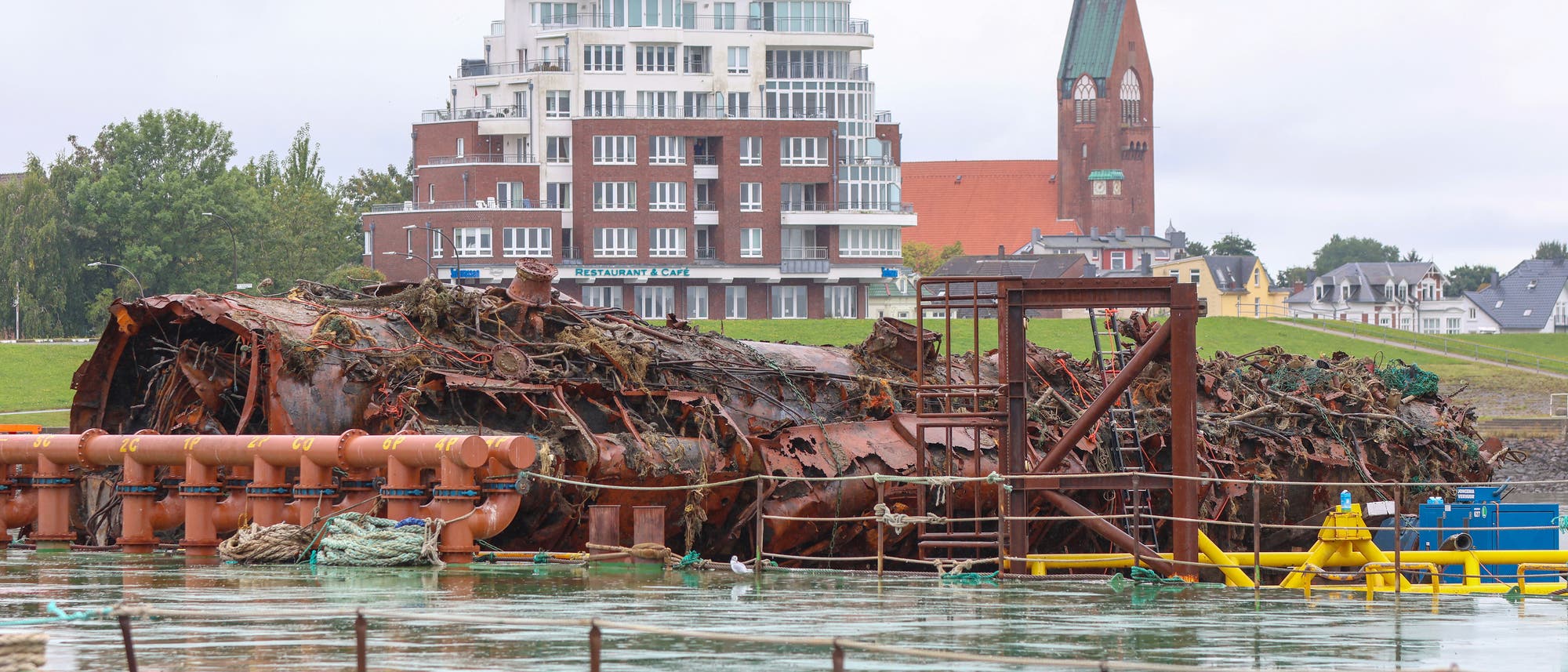 Ein altes, rostiges Schiffswrack liegt im Wasser vor einer modernen Wohnanlage und einem historischen Kirchturm. Im Vordergrund sind industrielle Rohre und Strukturen zu sehen. Die Szene zeigt einen Kontrast zwischen der verfallenen Struktur und der urbanen Umgebung im Hintergrund.
