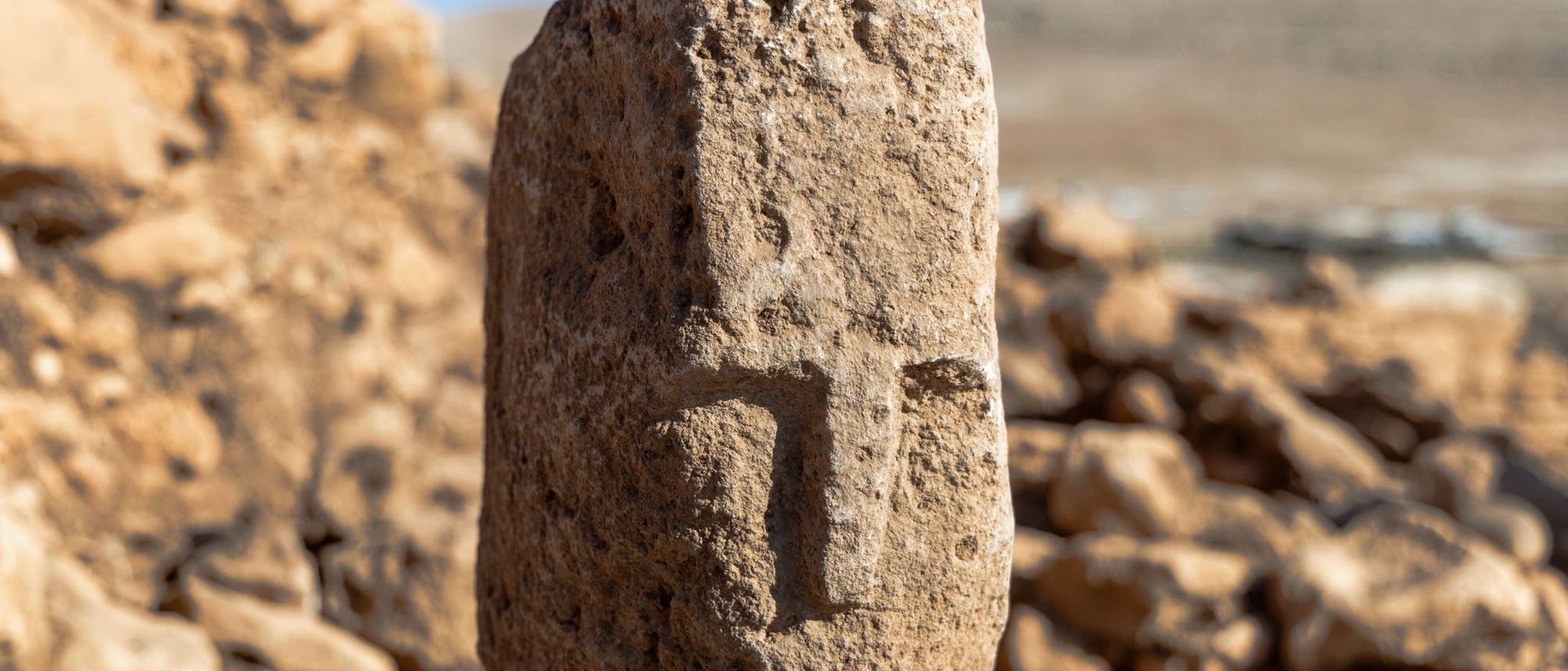 Ein alter, verwitterter Steinpfeiler mit eingraviertem, stark abstrahiertem Gesicht steht im Vordergrund einer archäologischen Ausgrabungsstätte. Der Hintergrund zeigt blauen Himmel und Landschaft. Der Fokus liegt auf den Details der Steinstruktur und der Gravur. Ein alter, verwitterter Steinpfeiler mit eingraviertem, stark abstrahiertem Gesicht steht im Vordergrund einer archäologischen Ausgrabungsstätte. Der Hintergrund zeigt blauen Himmel und Landschaft. Der Fokus liegt auf den Details der Steinstruktur und der Gravur.