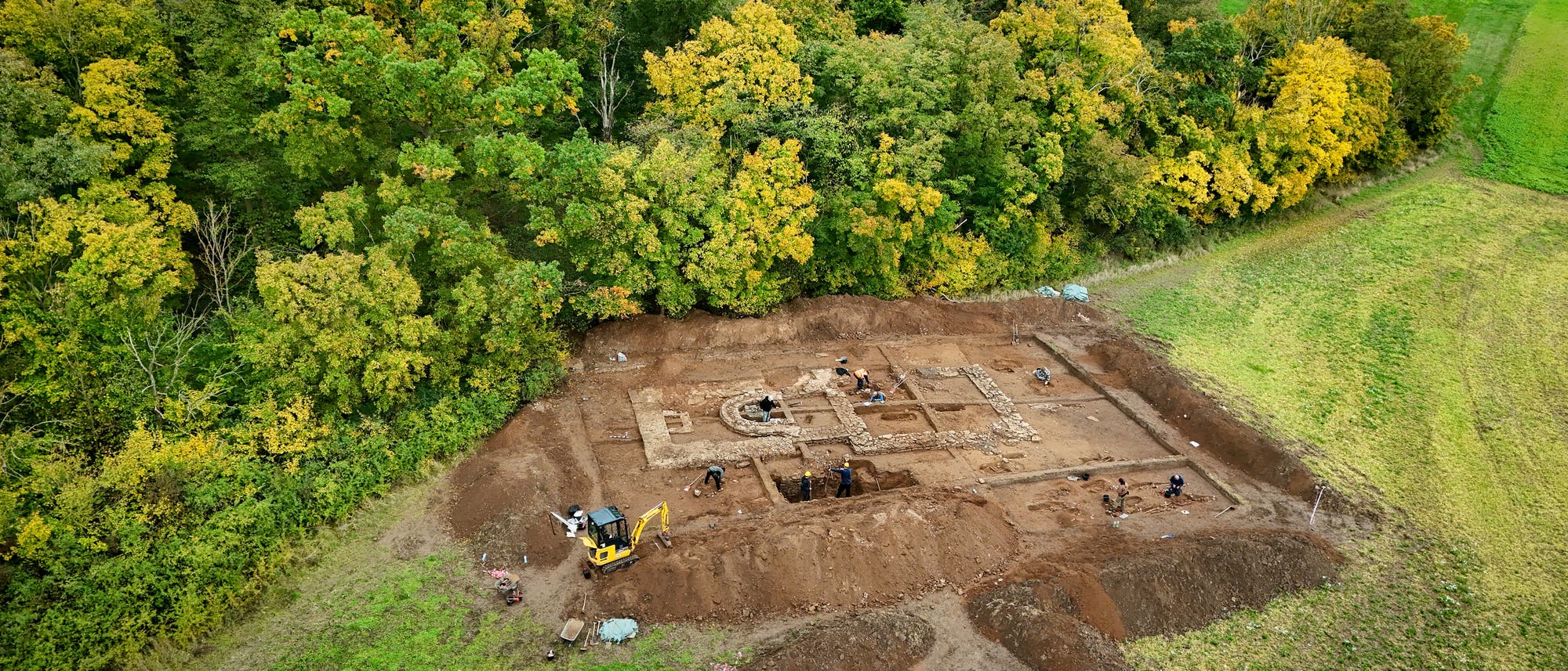 Luftaufnahme einer arch&auml;ologischen Ausgrabungsst&auml;tte am Waldrand. Mehrere Personen arbeiten an verschiedenen Stellen der Ausgrabung, die rechteckige Strukturen und freigelegte Mauerreste zeigt. Ein kleiner Bagger ist im Einsatz, um Erde zu bewegen. Die Umgebung ist von gr&uuml;nen und herbstlich gef&auml;rbten B&auml;umen sowie Wiesen umgeben.