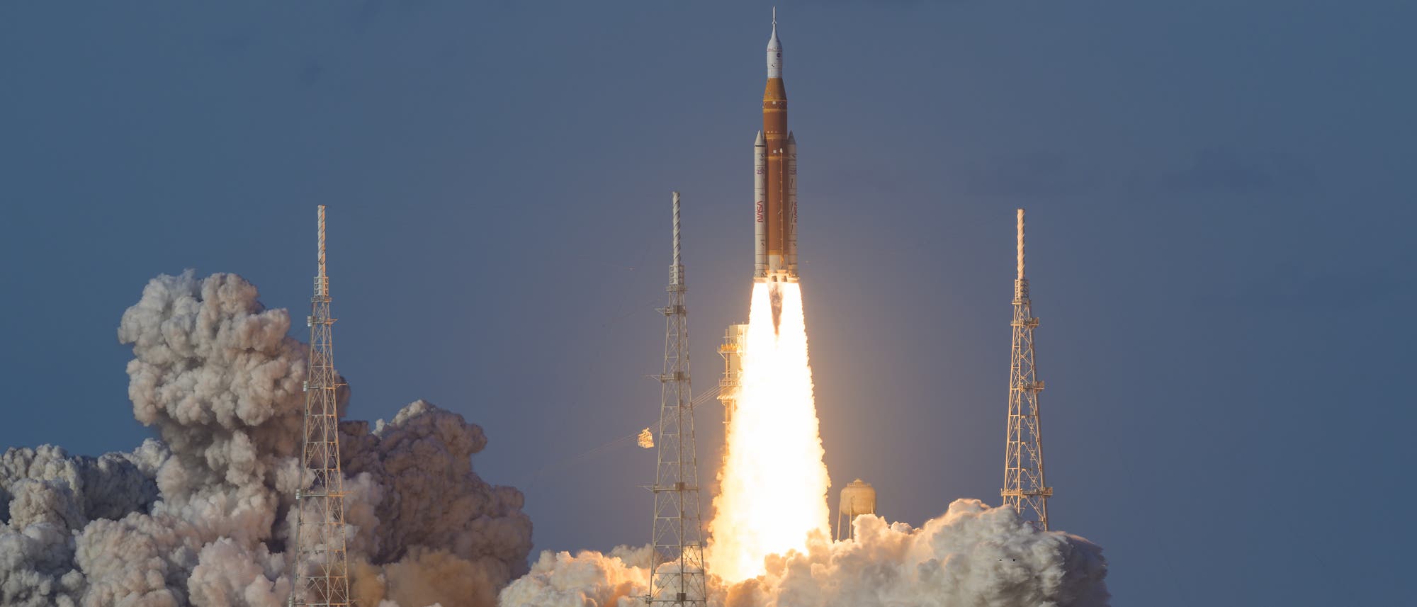 Eine Rakete hebt von einer Startrampe ab, umgeben von Rauch und Flammen. Der Himmel ist klar und blau. Die Rakete ist von mehreren T&uuml;rmen umgeben, die Teil der Startinfrastruktur sind.