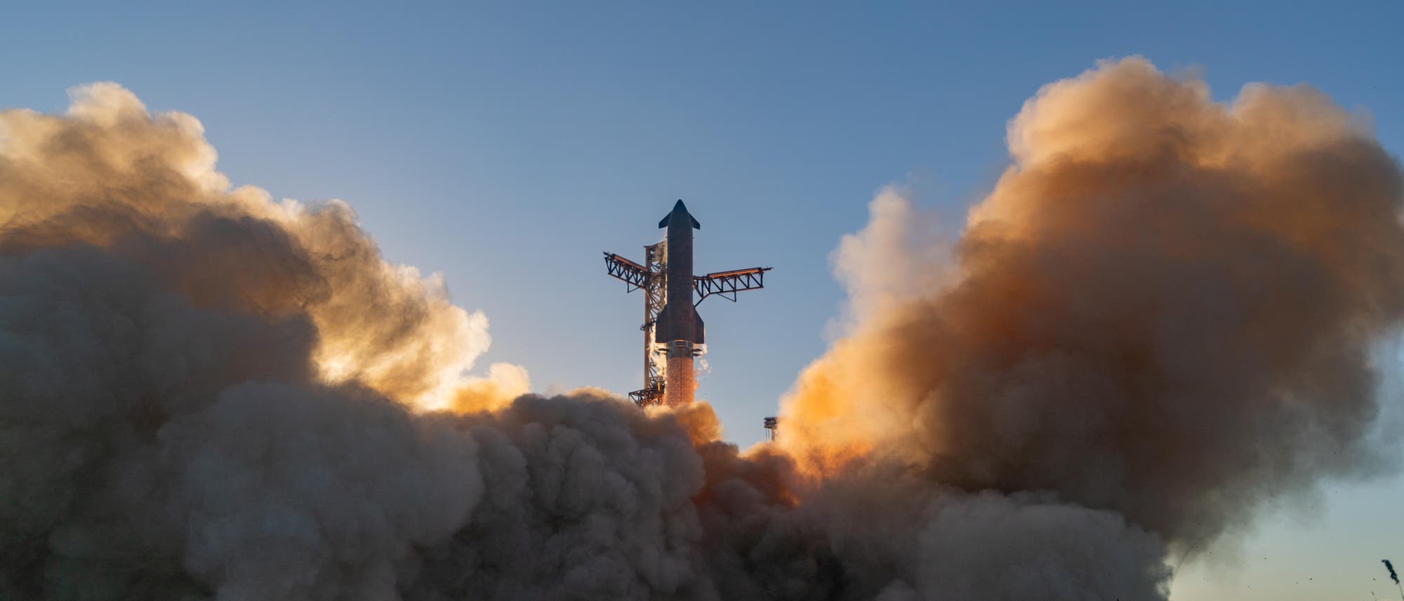 Eine Rakete hebt von einer Startrampe ab, umgeben von dichten Rauch- und Staubwolken. Der Himmel ist klar und blau, während die Sonne im Hintergrund die Szene beleuchtet. Die Rakete ist vertikal ausgerichtet und von metallischen Strukturen umgeben.