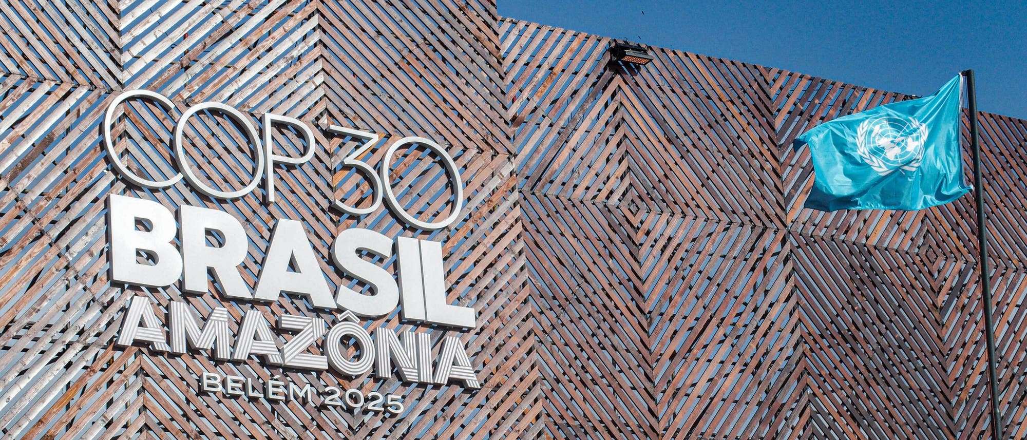 Gebäude mit einer modernen, geometrischen Fassade, auf der in großen Buchstaben "COP30 BRASIL AMAZÔNIA BELÉM 2025" steht. Rechts weht eine blaue Flagge mit dem Logo der Vereinten Nationen. Der Himmel ist klar und blau.
