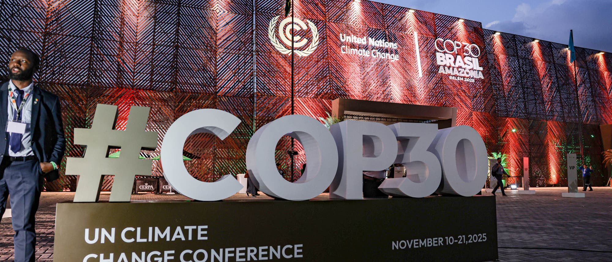 Eine große Skulptur mit der Aufschrift "#COP30" steht vor einem beleuchteten Gebäude, das für die UN-Klimakonferenz 2025 in Brasilien, Amazonas, vorbereitet ist. Auf dem Gebäude sind die Logos der Vereinten Nationen und der Konferenz sichtbar. Menschen gehen an der Szenerie vorbei. Der Himmel ist bewölkt, und die Umgebung ist in warmen Farben beleuchtet.