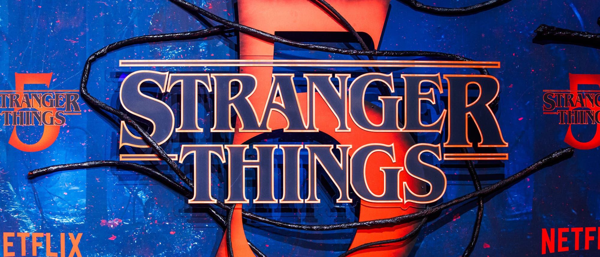 Ein k&uuml;nstlerisches Poster mit dem Titel "Stranger Things" in gro&szlig;en, auff&auml;lligen Buchstaben. Im Hintergrund ist eine gro&szlig;e rote Zahl 5 zu sehen, die von schwarzen, tentakelartigen Linien umgeben ist. Der Hintergrund ist in einem lebhaften Blau gehalten, mit mehreren "Netflix"-Logos in Orange. Das Design vermittelt ein mysteri&ouml;ses und spannendes Gef&uuml;hl, passend zur Serie.