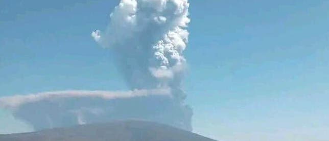 Ein Vulkan in einer kargen Landschaft stößt eine große, weiße Rauch- und Aschewolke in den klaren blauen Himmel aus. Die Wolke steigt hoch auf und breitet sich nach oben hin aus. Im Vordergrund ist eine flache, trockene Ebene zu sehen. Ein Vulkan in einer kargen Landschaft stößt eine große, weiße Rauch- und Aschewolke in den klaren blauen Himmel aus. Die Wolke steigt hoch auf und breitet sich nach oben hin aus. Im Vordergrund ist eine flache, trockene Ebene zu sehen.
