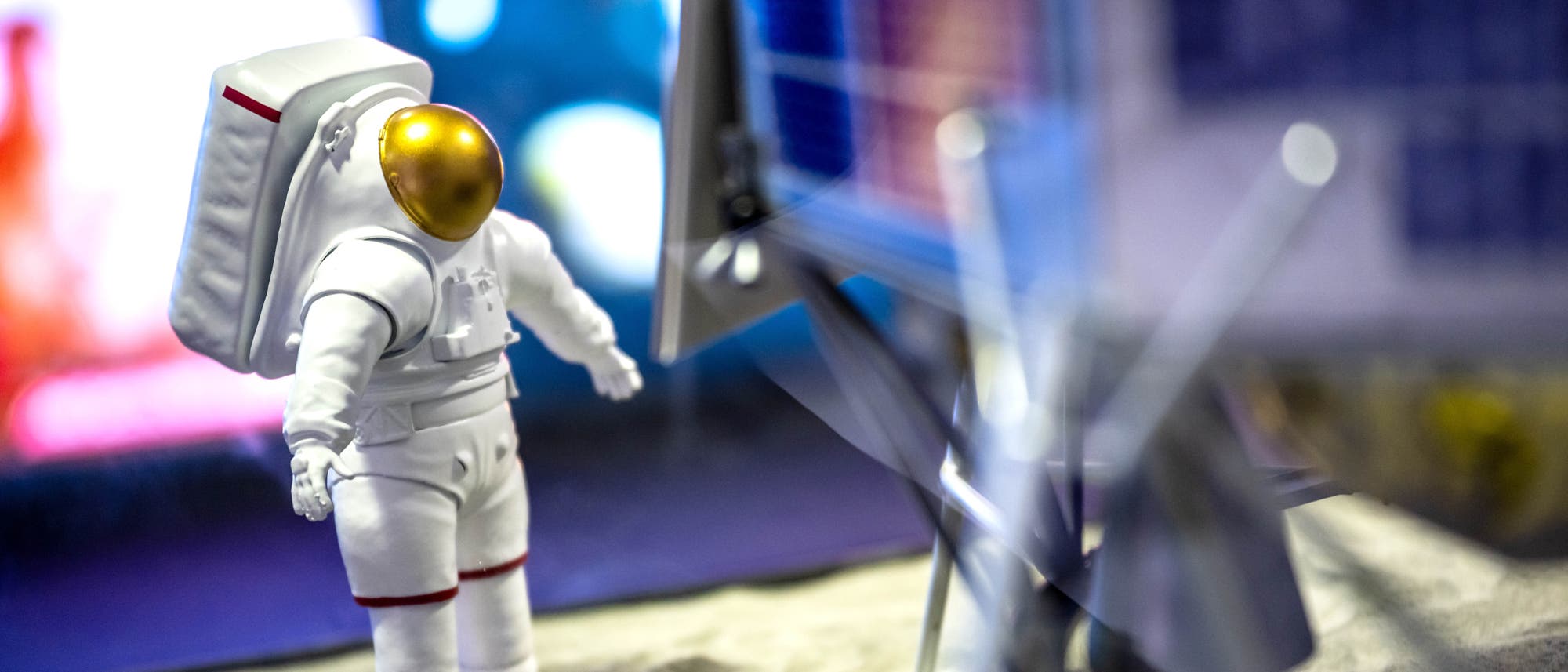 Die Kunststofffigur eines Astronauten in einem wei&szlig;en Raumanzug mit goldfarbenem Visier steht auf einer sandigen Oberfl&auml;che neben dem Modell eines Raumfahrzeugs. Der Hintergrund ist unscharf und zeigt bunte Lichter, die eine futuristische oder au&szlig;erirdische Umgebung andeuten. Der Astronaut scheint das Raumfahrzeug zu inspizieren.