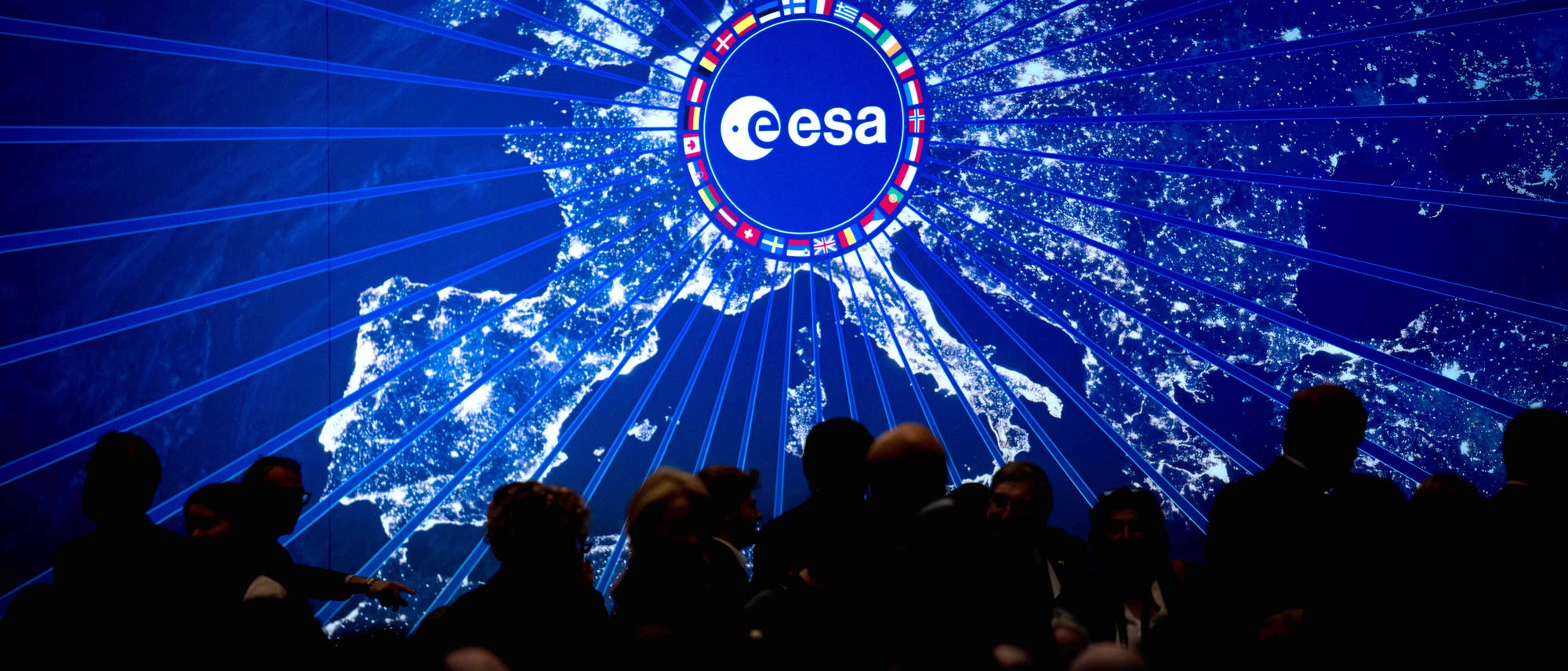 Eine nächtliche Satellitenaufnahme von Europa, die von einem zentralen Punkt aus strahlenförmig beleuchtet wird. Im Zentrum befindet sich das Logo der Europäischen Weltraumorganisation (ESA), umgeben von Flaggen verschiedener Länder. Im Vordergrund sind die Silhouetten von Menschen zu sehen, die die Karte betrachten. Die Darstellung hebt die Vernetzung und Zusammenarbeit der europäischen Länder im Bereich der Raumfahrt hervor. Eine nächtliche Satellitenaufnahme von Europa, die von einem zentralen Punkt aus strahlenförmig beleuchtet wird. Im Zentrum befindet sich das Logo der Europäischen Weltraumorganisation (ESA), umgeben von Flaggen verschiedener Länder. Im Vordergrund sind die Silhouetten von Menschen zu sehen, die die Karte betrachten. Die Darstellung hebt die Vernetzung und Zusammenarbeit der europäischen Länder im Bereich der Raumfahrt hervor.