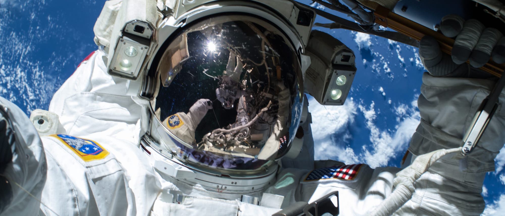 Ein Astronaut in einem wei&szlig;en Raumanzug f&uuml;hrt einen Weltraumspaziergang durch. Der Helm spiegelt die Erde und Teile der Raumstation wider. Im Hintergrund ist der blaue Planet mit wei&szlig;en Wolkenformationen zu sehen. Der Astronaut h&auml;lt sich an einer Struktur der Raumstation fest. Auf dem Anzug ist eine Flagge zu erkennen.