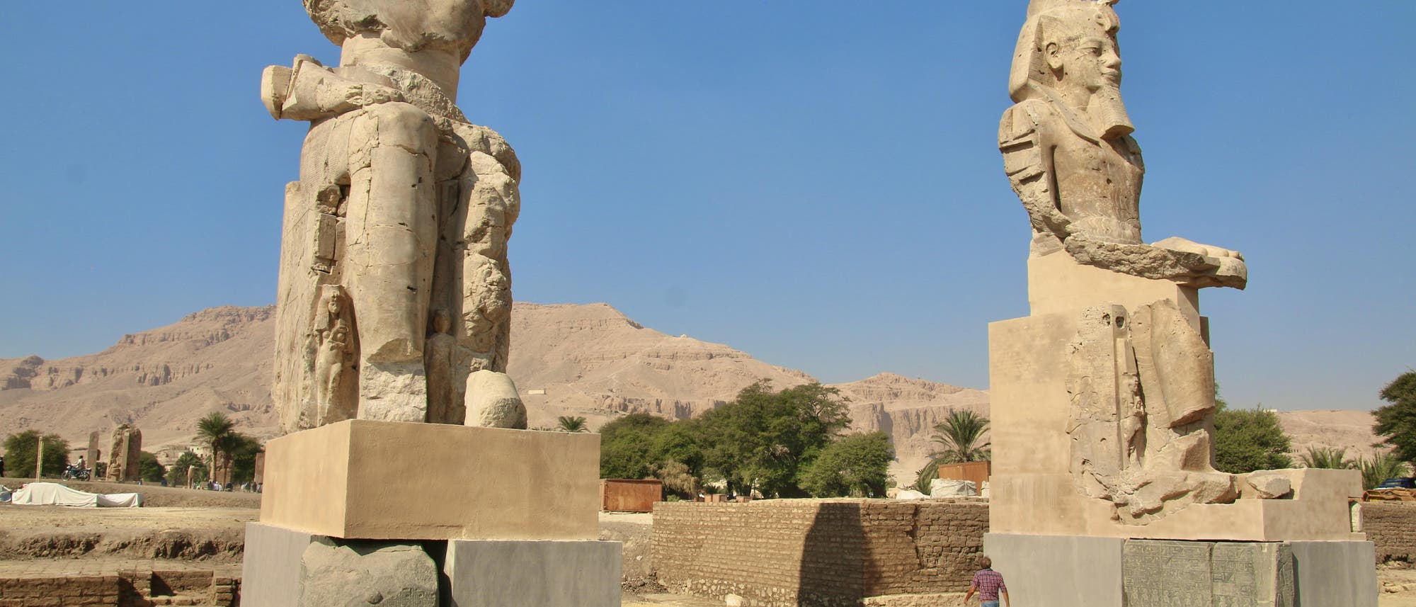 Zwei gro&szlig;e, besch&auml;digte Sitzstatuen aus Stein stehen in einer W&uuml;stenlandschaft mit Bergen im Hintergrund. Eine Person geht an der rechten Statue vorbei, was die Gr&ouml;&szlig;e der Monumente verdeutlicht. Der Himmel ist klar und blau.