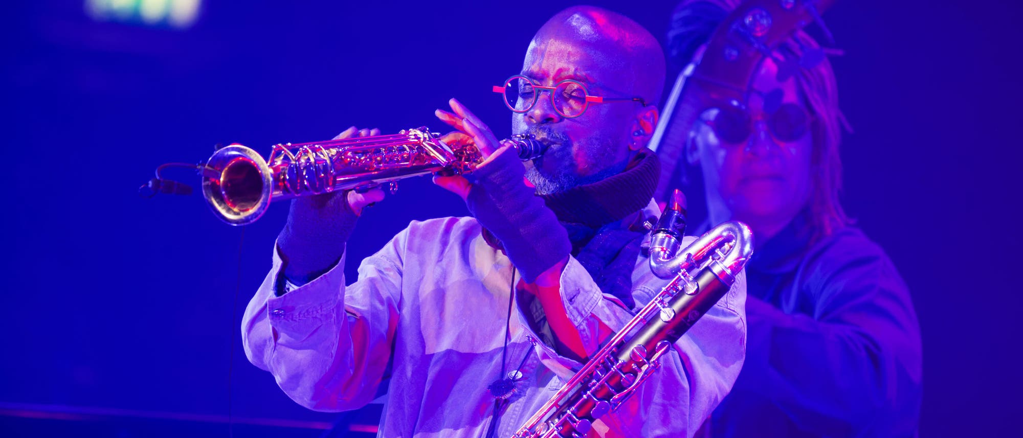 Zwei Musiker spielen auf einer B&uuml;hne in blauer Beleuchtung. Der vordere Musiker spielt ein Saxophon, w&auml;hrend der hintere Musiker einen Bass h&auml;lt. Beide tragen Brillen und sind in konzentrierter Haltung. Die Szene vermittelt eine lebendige und dynamische Atmosph&auml;re.