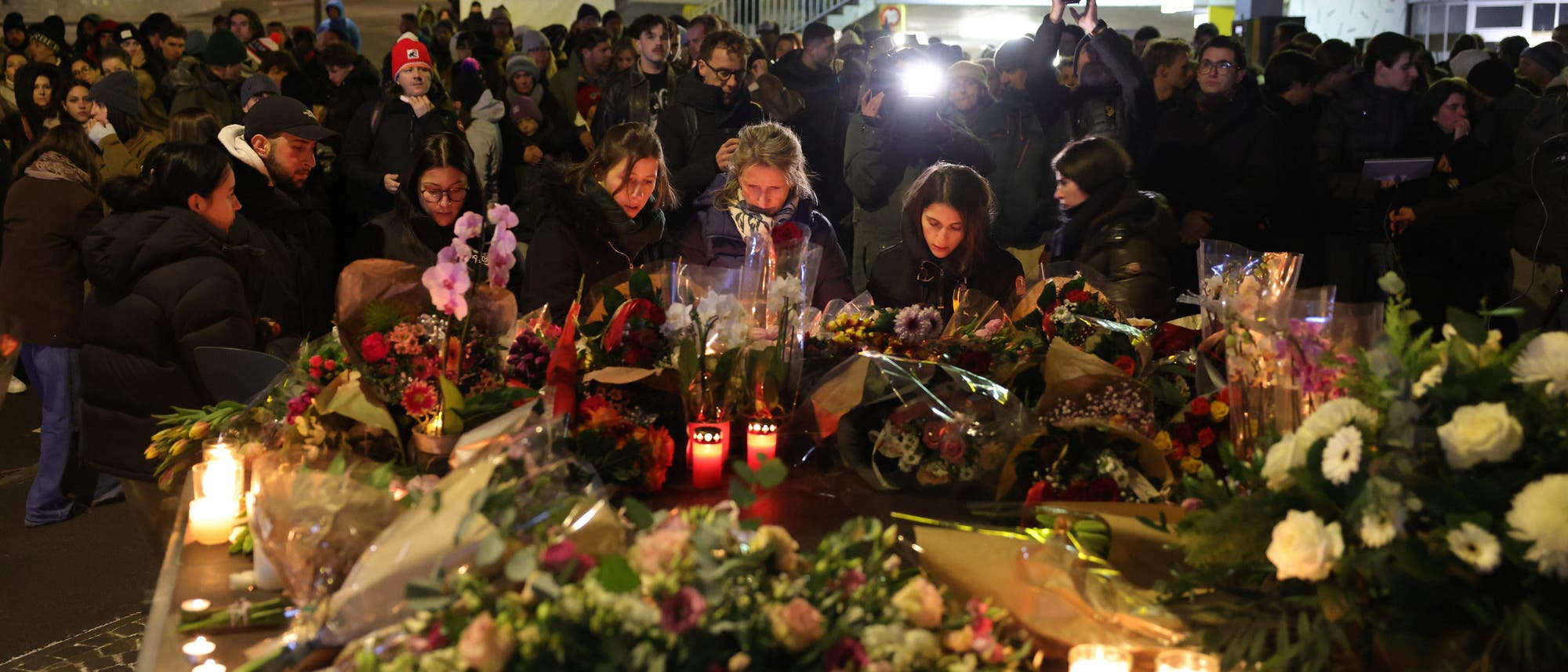 Eine Menschenmenge versammelt sich bei Nacht um einen improvisierten Gedenkort mit Blumen und Kerzen. Einige Personen stehen nahe beieinander und betrachten die Blumenarrangements. Im Hintergrund sind weitere Menschen zu sehen, einige mit Kameras. Die Szene ist von ged&auml;mpftem Licht und einer ruhigen, nachdenklichen Atmosph&auml;re gepr&auml;gt.
