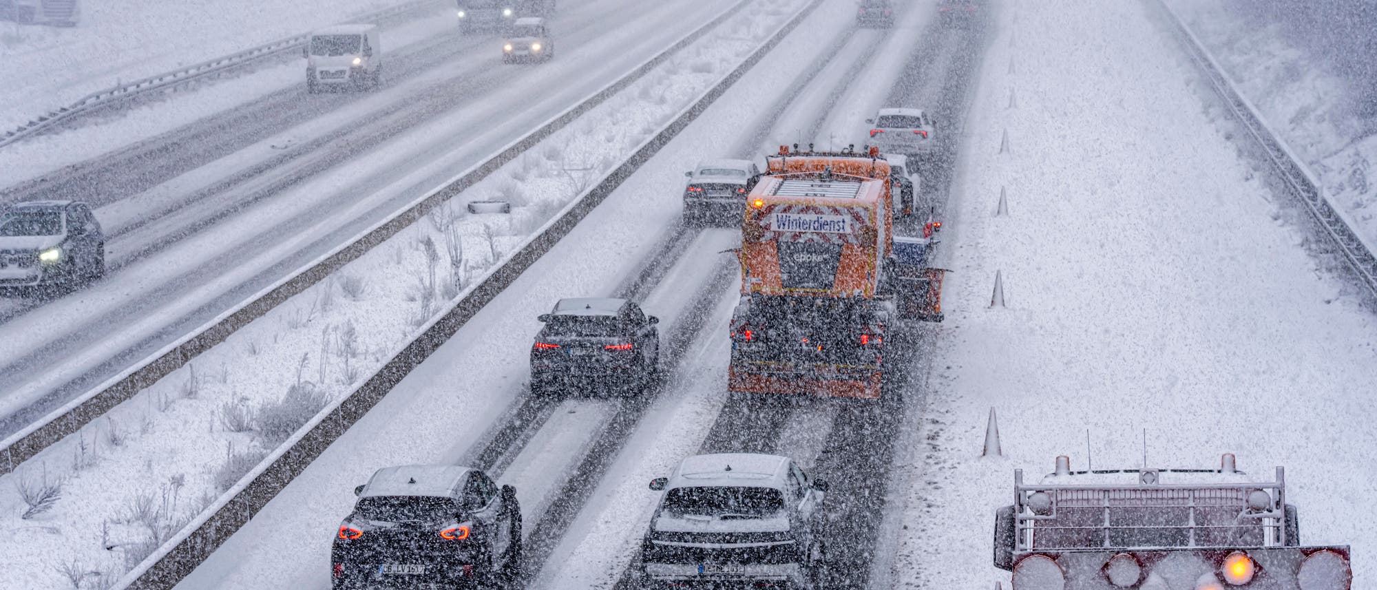 Eine verschneite Autobahn mit mehreren Fahrspuren, auf der Autos und ein Schneepflug fahren. Der Schneepflug ist mit dem Schriftzug "Winterdienst" gekennzeichnet. Die Straße ist mit einer dünnen Schneeschicht bedeckt, und es schneit stark. Die Sicht ist eingeschränkt, und die Fahrzeuge fahren vorsichtig. Im Vordergrund ist ein beleuchtetes Warnschild zu sehen. Eine verschneite Autobahn mit mehreren Fahrspuren, auf der Autos und ein Schneepflug fahren. Der Schneepflug ist mit dem Schriftzug "Winterdienst" gekennzeichnet. Die Straße ist mit einer dünnen Schneeschicht bedeckt, und es schneit stark. Die Sicht ist eingeschränkt, und die Fahrzeuge fahren vorsichtig. Im Vordergrund ist ein beleuchtetes Warnschild zu sehen.