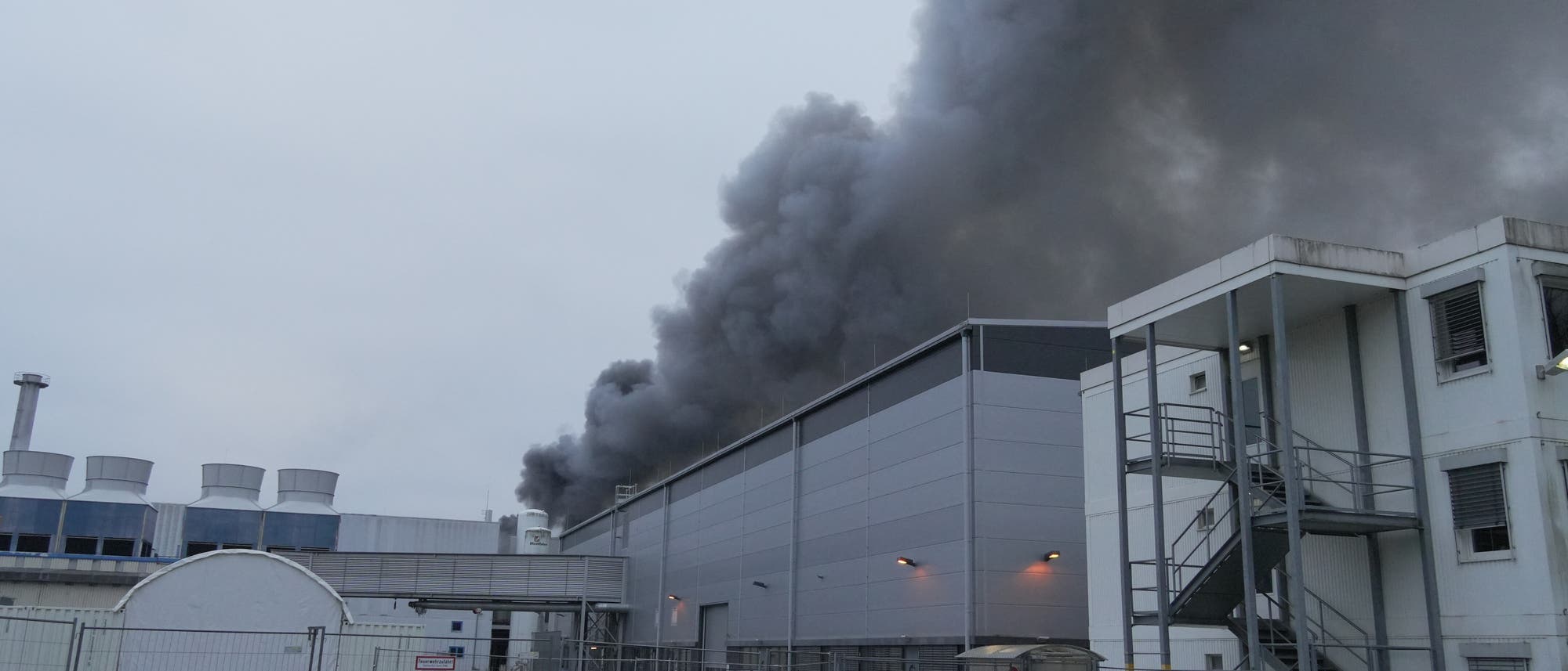 Eine Industrieanlage mit grauen Geb&auml;uden, aus der dichter, dunkler Rauch aufsteigt, was auf einen Brand hindeutet. Im Vordergrund sind ein Zaun und ein Parkplatz mit einem Auto zu sehen. Der Himmel ist bew&ouml;lkt, und die Szene wirkt k&uuml;hl und d&uuml;ster.
