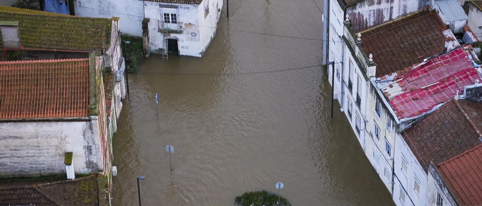 Luftaufnahme einer &uuml;berfluteten Stadtstra&szlig;e. Mehrere Geb&auml;ude mit roten Ziegeld&auml;chern sind von braunem Hochwasser umgeben. Stra&szlig;enschilder und Laternenmasten ragen aus dem Wasser. Ein kleines Boot ist am oberen Bildrand zu sehen. Die Szene zeigt die Auswirkungen einer &Uuml;berschwemmung auf eine st&auml;dtische Umgebung.