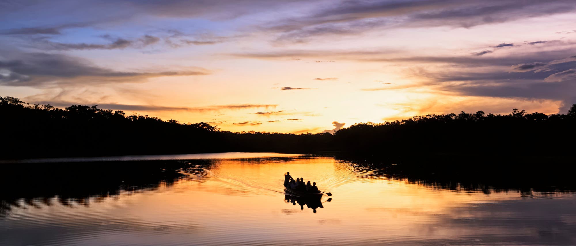 Eine Gruppe von Menschen f&auml;hrt in einem kleinen Boot &uuml;ber einen ruhigen See bei Sonnenuntergang. Der Himmel ist in warmen Gelb- und Oranget&ouml;nen gef&auml;rbt, w&auml;hrend sich die Silhouetten der B&auml;ume am Horizont abzeichnen. Die Wasseroberfl&auml;che spiegelt die Farben des Himmels wider und verst&auml;rkt die friedliche Atmosph&auml;re der Szene.