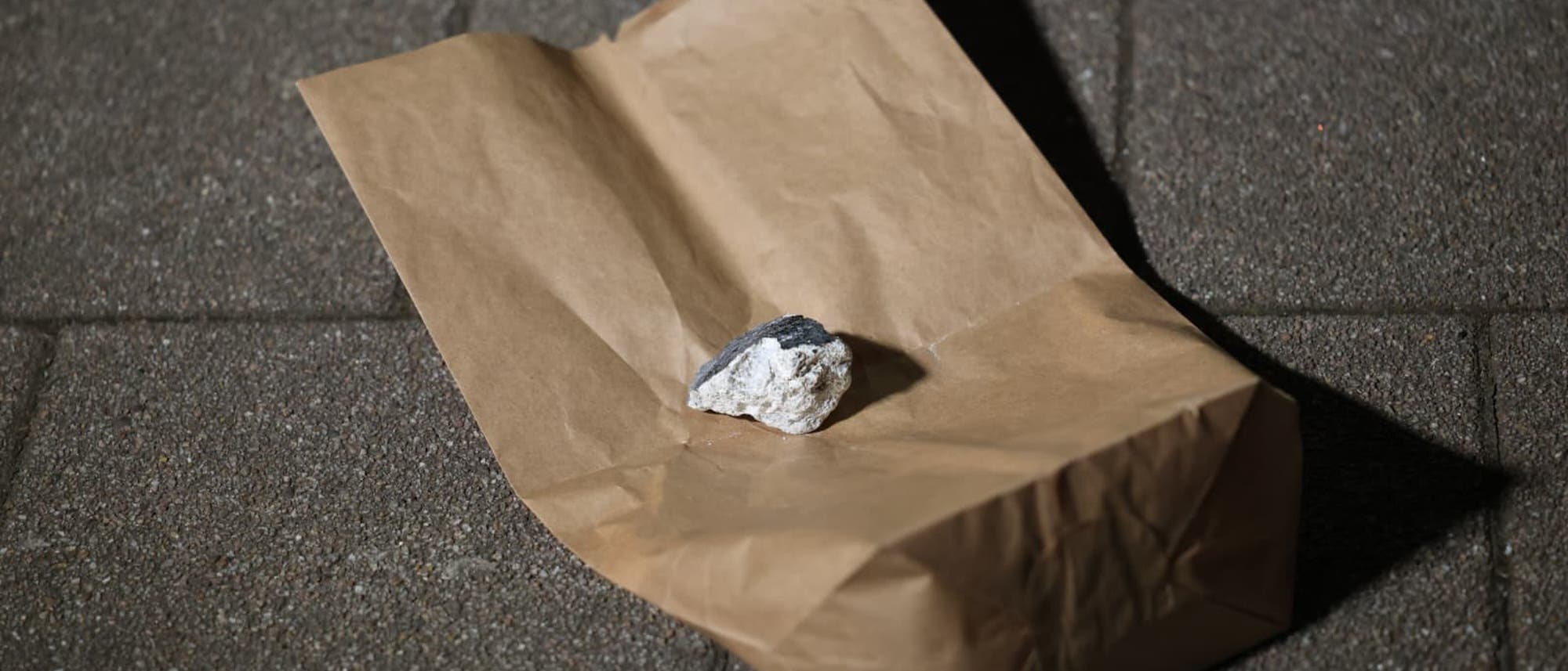 Eine braune Papiert&uuml;te liegt auf einem gepflasterten Boden. Auf der T&uuml;te befindet sich ein kleiner, unregelm&auml;&szlig;ig geformter Stein. Die Szene ist bei schwachem Licht aufgenommen, was Schatten auf dem Boden erzeugt.
