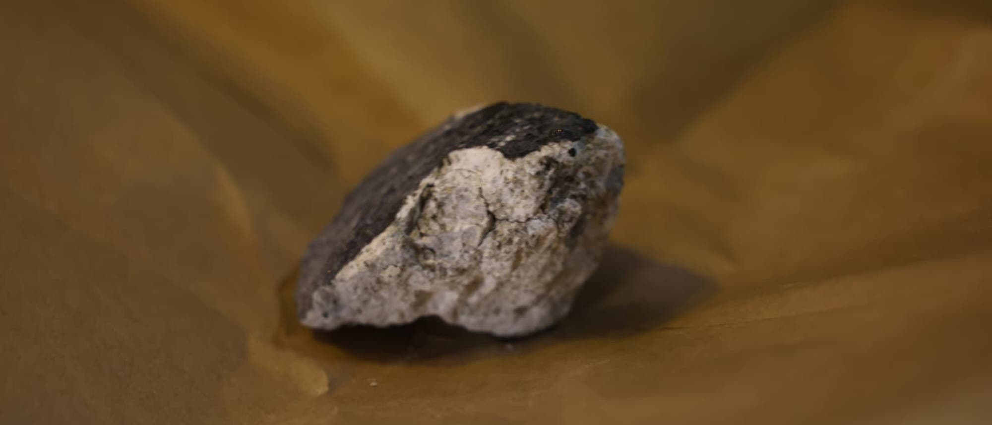 Ein grober, unregelmäßig geformter Stein liegt auf braunem Papier. Der Stein hat eine dunkle, raue Oberfläche mit helleren, glatten Bereichen. Die Szene ist in warmen, erdigen Tönen gehalten. Ein grober, unregelmäßig geformter Stein liegt auf braunem Papier. Der Stein hat eine dunkle, raue Oberfläche mit helleren, glatten Bereichen. Die Szene ist in warmen, erdigen Tönen gehalten.