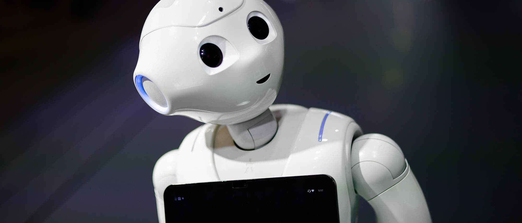 Ein humanoider Roboter mit einem weißen, glatten Gehäuse und großen, runden Augen steht in einem Ausstellungsraum. Der Roboter trägt ein Tablet auf der Brust, auf dem Text angezeigt wird. Der Hintergrund ist unscharf, was den Fokus auf den Roboter lenkt. Der Roboter scheint freundlich zu wirken und ist leicht geneigt, als ob er zuhört. Ein humanoider Roboter mit einem weißen, glatten Gehäuse und großen, runden Augen steht in einem Ausstellungsraum. Der Roboter trägt ein Tablet auf der Brust, auf dem Text angezeigt wird. Der Hintergrund ist unscharf, was den Fokus auf den Roboter lenkt. Der Roboter scheint freundlich zu wirken und ist leicht geneigt, als ob er zuhört.