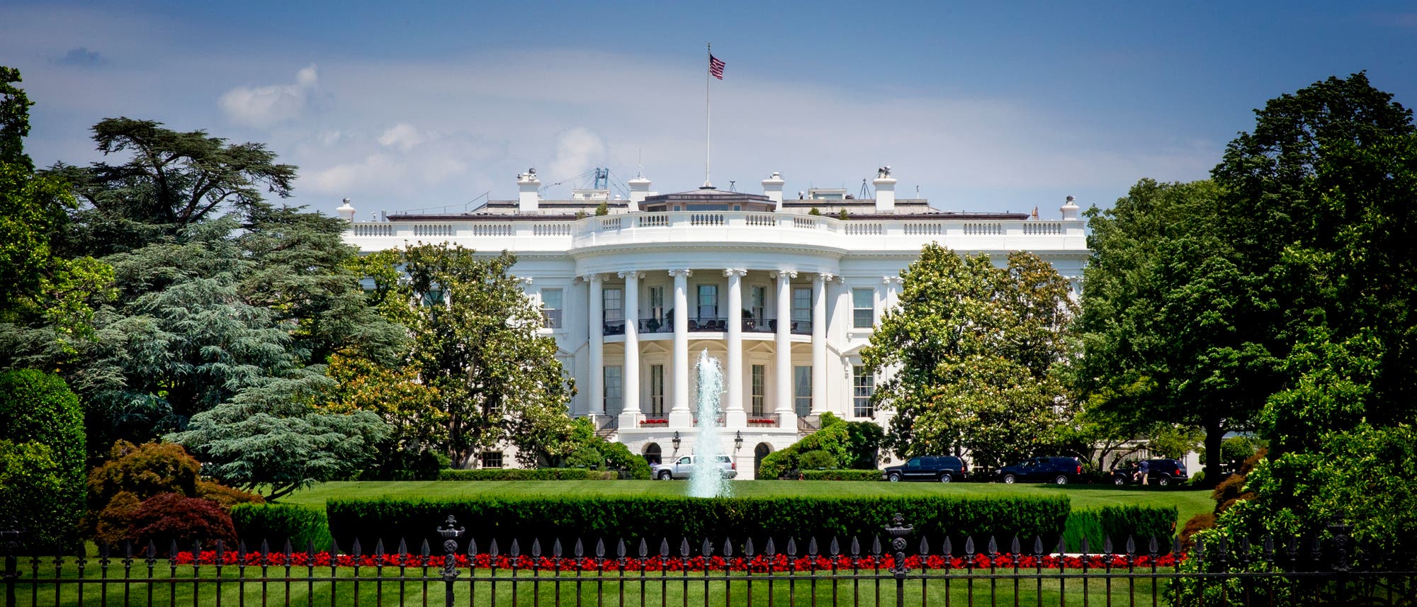 Das Bild zeigt das Weiße Haus in Washington, D.C., ein ikonisches Gebäude mit einer weißen Fassade und Säulen, umgeben von gepflegten Rasenflächen und Bäumen. Eine amerikanische Flagge weht auf dem Dach. Der Himmel ist blau mit einigen Wolken.