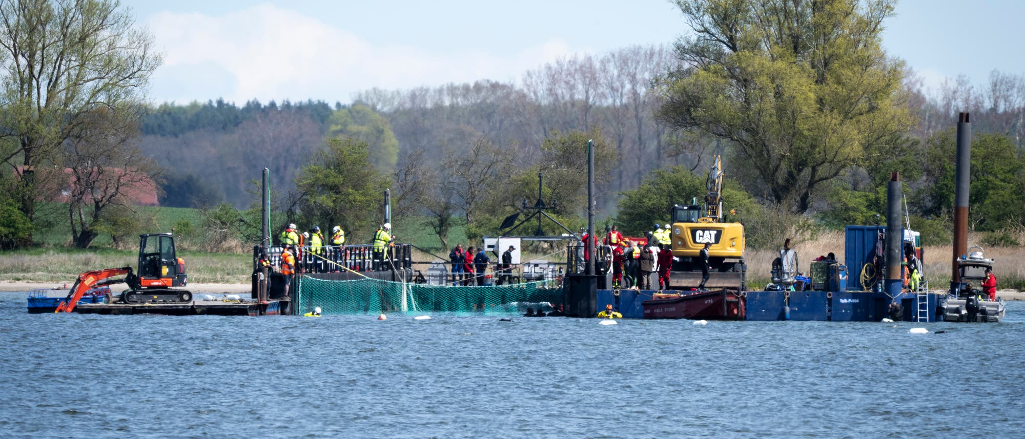 Eine Gruppe von Menschen arbeitet auf einer schwimmenden Plattform auf einem Gew&auml;sser. Die Plattform ist mit Bauger&auml;ten wie einem Bagger und einem Kran ausgestattet. Personen in Sicherheitskleidung sind mit verschiedenen Aufgaben besch&auml;ftigt. Im Hintergrund sind B&auml;ume und eine bewaldete Uferlandschaft zu sehen. Das Bild zeigt eine Baustelle auf dem Wasser, m&ouml;glicherweise f&uuml;r ein Bau- oder Infrastrukturprojekt.