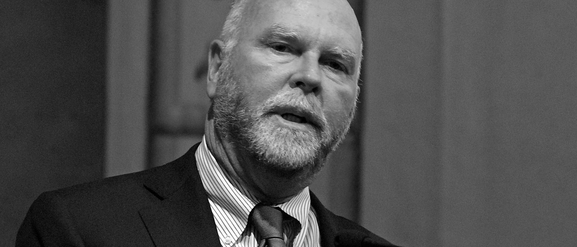 Ein Mann in einem Anzug spricht in ein Mikrofon. Der Hintergrund ist unscharf, und das Bild ist in Schwarz-Wei&szlig; gehalten.