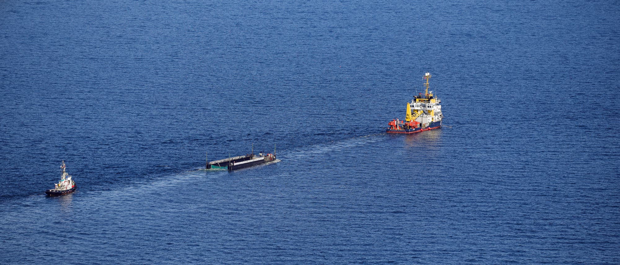 Ein gro&szlig;es Schiff zieht ein l&auml;ngliches Objekt &uuml;ber das offene Meer, begleitet von einem kleineren Boot. Die Szene zeigt eine ruhige Wasseroberfl&auml;che unter einem klaren Himmel. Das Hauptschiff ist bunt bemalt und scheint f&uuml;r spezielle Aufgaben ausger&uuml;stet zu sein.