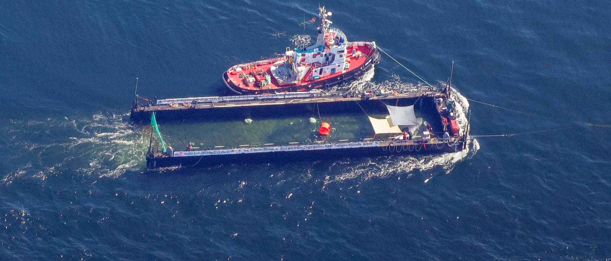 Luftaufnahme eines roten Schleppers, der einen langen, rechteckigen Lastkahn auf dem Meer zieht. Auf dem Flo&szlig; befinden sich mehrere Ausr&uuml;stungsgegenst&auml;nde, darunter ein gro&szlig;es Netz und orangefarbene Bojen. Das Wasser um das Flo&szlig; ist leicht aufgew&uuml;hlt.