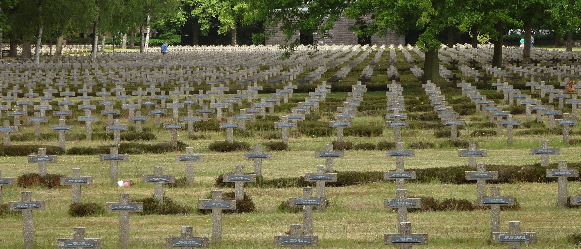 Ein weitläufiger Soldatenfriedhof mit zahlreichen grauen Steinkreuzen, die in geordneten Reihen aufgestellt sind. Im Hintergrund sind grüne Bäume zu sehen. Eine einzelne Person in blauer Kleidung geht zwischen den Gräbern entlang. Die Szene vermittelt eine ruhige und nachdenkliche Atmosphäre. Ein weitläufiger Soldatenfriedhof mit zahlreichen grauen Steinkreuzen, die in geordneten Reihen aufgestellt sind. Im Hintergrund sind grüne Bäume zu sehen. Eine einzelne Person in blauer Kleidung geht zwischen den Gräbern entlang. Die Szene vermittelt eine ruhige und nachdenkliche Atmosphäre.