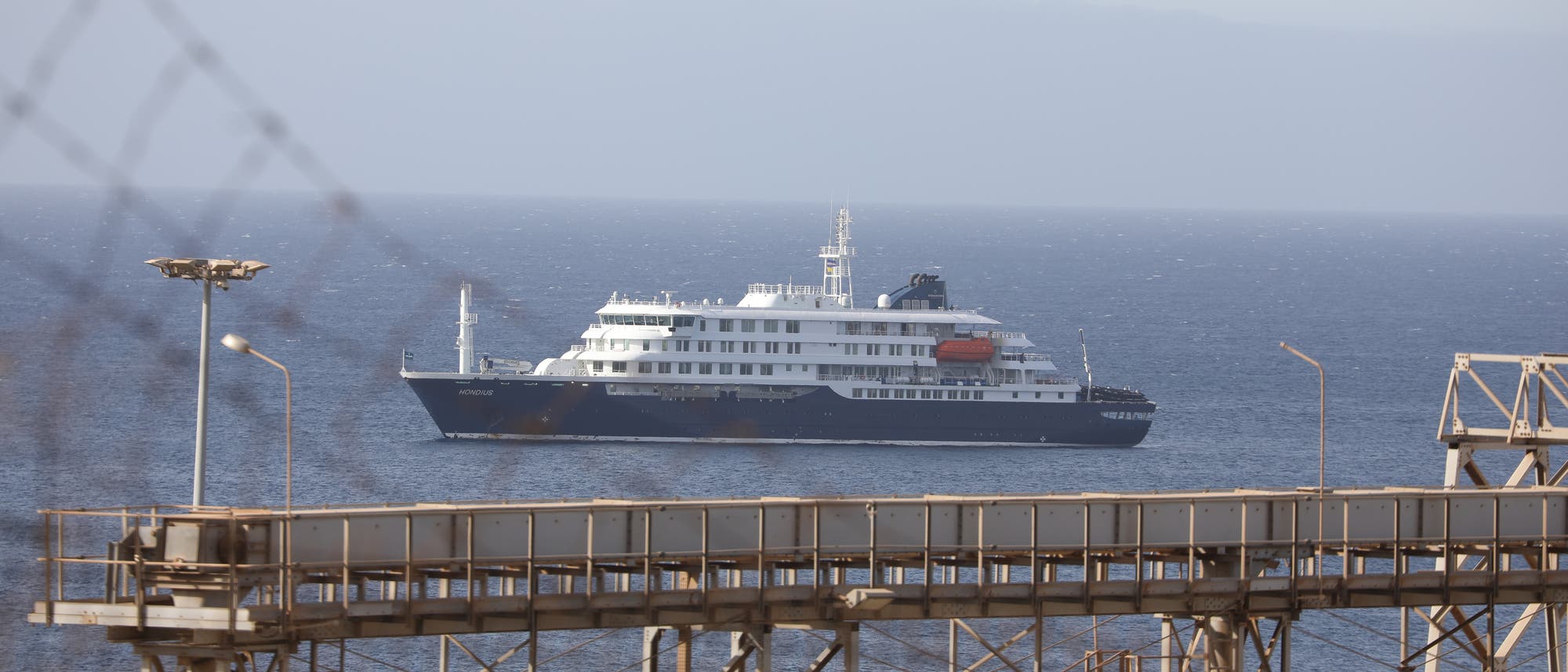 Ein gro&szlig;es Kreuzfahrtschiff f&auml;hrt auf einem ruhigen Meer, im Hintergrund sind verschwommene Berge zu sehen. Im Vordergrund befinden sich industrielle Strukturen und ein Lichtmast. Das Schiff ist wei&szlig; mit blauen Akzenten und hat mehrere Decks.