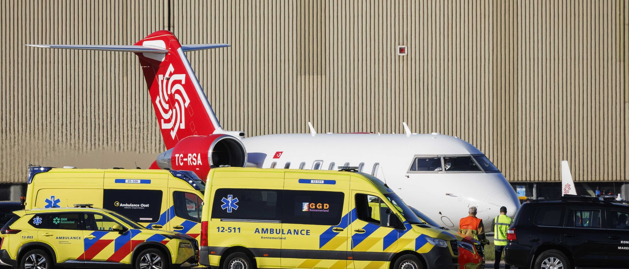Ein Flugzeug steht auf einem Flughafen vor einem gro&szlig;en Hangar. Im Vordergrund sind zwei gelbe Krankenwagen mit blauen und roten Streifen zu sehen, die mit "Ambulance" und "GGD" beschriftet sind. Zwei Personen in Sicherheitswesten stehen neben einem schwarzen Fahrzeug. Die Szene deutet auf einen medizinischen Einsatz oder eine Notfallsituation hin.