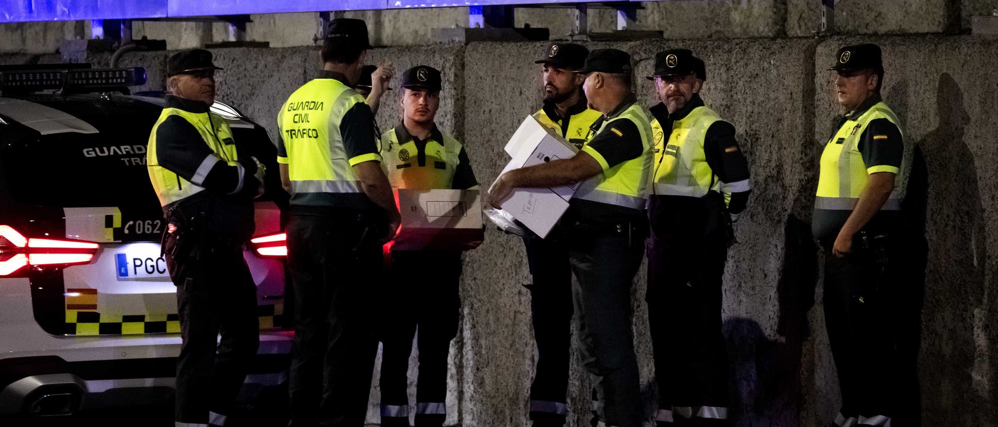 Eine Gruppe von Polizisten steht in einer nächtlichen Szene neben einem Polizeifahrzeug. Sie tragen reflektierende Westen mit der Aufschrift "Guardia Civil Tráfico" und diskutieren über Dokumente, die einer von ihnen in der Hand hält. Die Umgebung ist dunkel, und das Fahrzeug hat eingeschaltete Rücklichter. Die Szene vermittelt eine Atmosphäre von Zusammenarbeit und Einsatzbereitschaft. Eine Gruppe von Polizisten steht in einer nächtlichen Szene neben einem Polizeifahrzeug. Sie tragen reflektierende Westen mit der Aufschrift "Guardia Civil Tráfico" und diskutieren über Dokumente, die einer von ihnen in der Hand hält. Die Umgebung ist dunkel, und das Fahrzeug hat eingeschaltete Rücklichter. Die Szene vermittelt eine Atmosphäre von Zusammenarbeit und Einsatzbereitschaft.