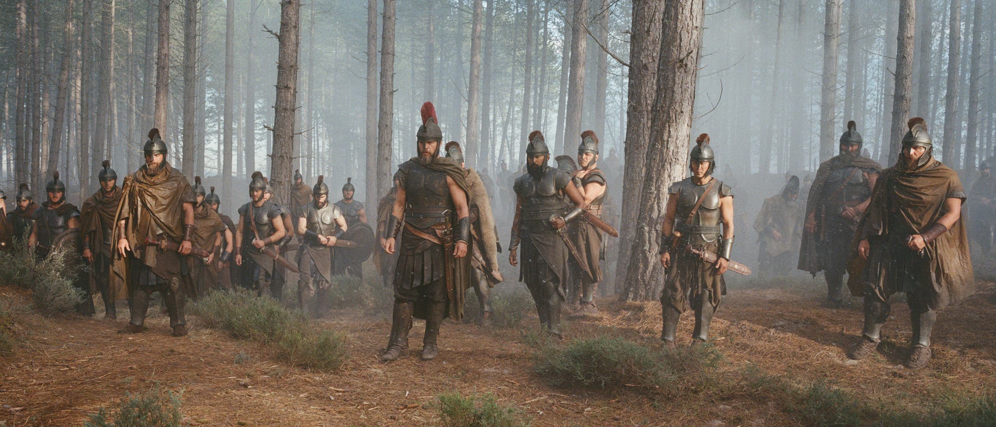 Eine Gruppe von Menschen in antiker Rüstung bewegt sich durch einen nebligen Wald. Sie tragen Helme und Umhänge und sind mit Schwertern bewaffnet. Die Szene vermittelt eine historische oder filmische Atmosphäre, die an eine militärische Expedition erinnert. Die Bäume sind hoch und der Boden ist mit Nadeln bedeckt. Eine Gruppe von Menschen in antiker Rüstung bewegt sich durch einen nebligen Wald. Sie tragen Helme und Umhänge und sind mit Schwertern bewaffnet. Die Szene vermittelt eine historische oder filmische Atmosphäre, die an eine militärische Expedition erinnert. Die Bäume sind hoch und der Boden ist mit Nadeln bedeckt.