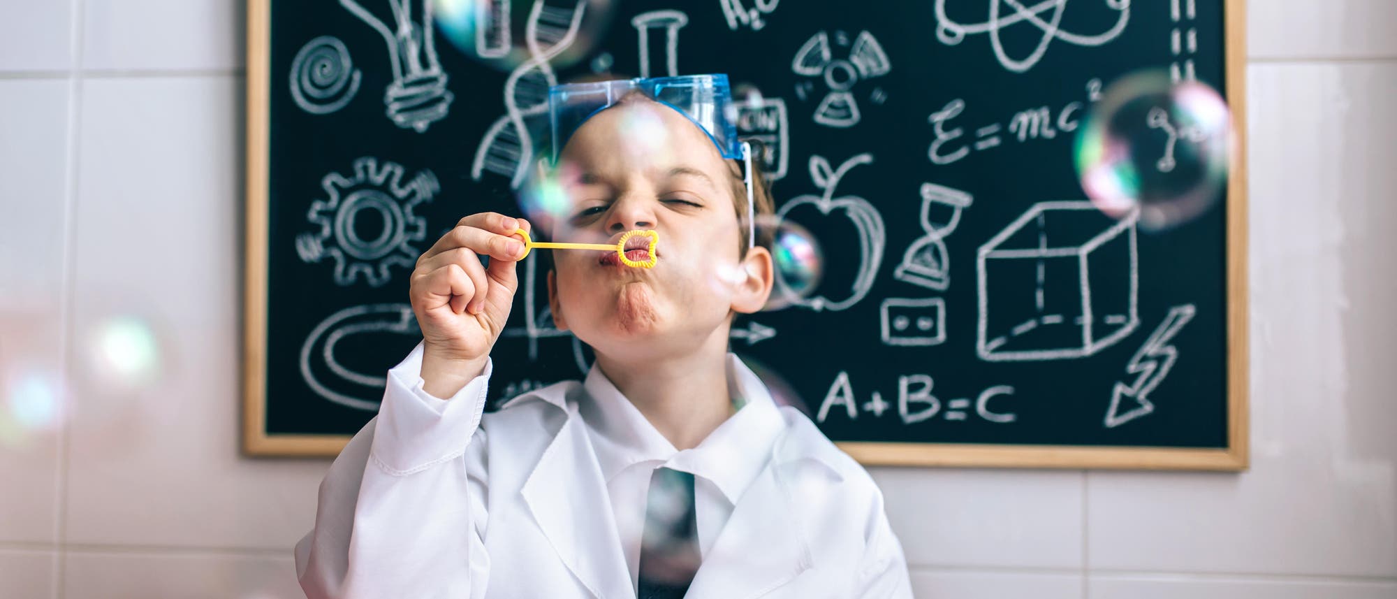 Ein Kind in einem wei&szlig;en Laborkittel bl&auml;st Seifenblasen. Im Hintergrund ist eine Tafel mit wissenschaftlichen Zeichnungen und Formeln wie E=mc&sup2; und A+B=C zu sehen. Das Kind tr&auml;gt eine Schutzbrille und wirkt konzentriert. Die Szene vermittelt eine spielerische Ann&auml;herung an Wissenschaft und Experimentieren.