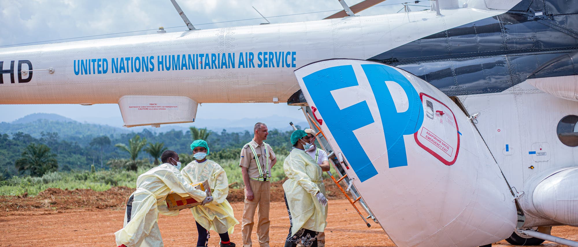 Eine Gruppe von Personen in Schutzkleidung entlädt Hilfsgüter aus einem Hubschrauber des United Nations Humanitarian Air Service. Der Hubschrauber steht auf einer unbefestigten Landebahn in einer ländlichen Umgebung mit bewaldeten Hügeln im Hintergrund. Die Personen tragen Gesichtsmasken und arbeiten zusammen, um die Lieferung effizient zu organisieren. Eine Gruppe von Personen in Schutzkleidung entlädt Hilfsgüter aus einem Hubschrauber des United Nations Humanitarian Air Service. Der Hubschrauber steht auf einer unbefestigten Landebahn in einer ländlichen Umgebung mit bewaldeten Hügeln im Hintergrund. Die Personen tragen Gesichtsmasken und arbeiten zusammen, um die Lieferung effizient zu organisieren.