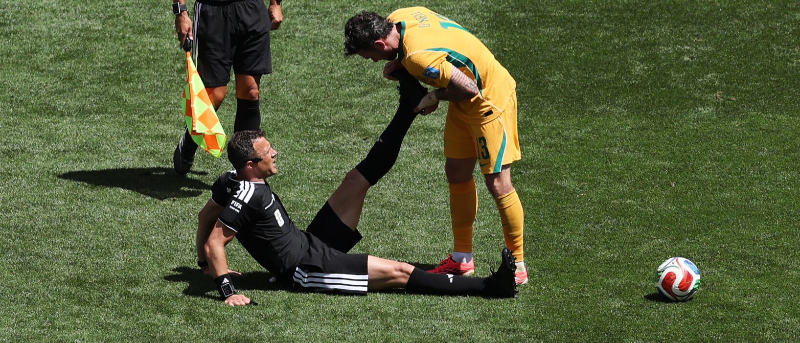 Der Fu&szlig;ballspieler  Aiden O&rsquo;Neill in einem gelben Trikot hilft Schiedsrichter Felix Zwayer, der auf dem Rasen sitzt, indem er dessen Bein dehnt. Ein weiterer Schiedsrichter steht daneben und beobachtet die Szene. Ein Fu&szlig;ball liegt in der N&auml;he auf dem Spielfeld. Die Szene findet auf einem gr&uuml;nen Fu&szlig;ballfeld statt.